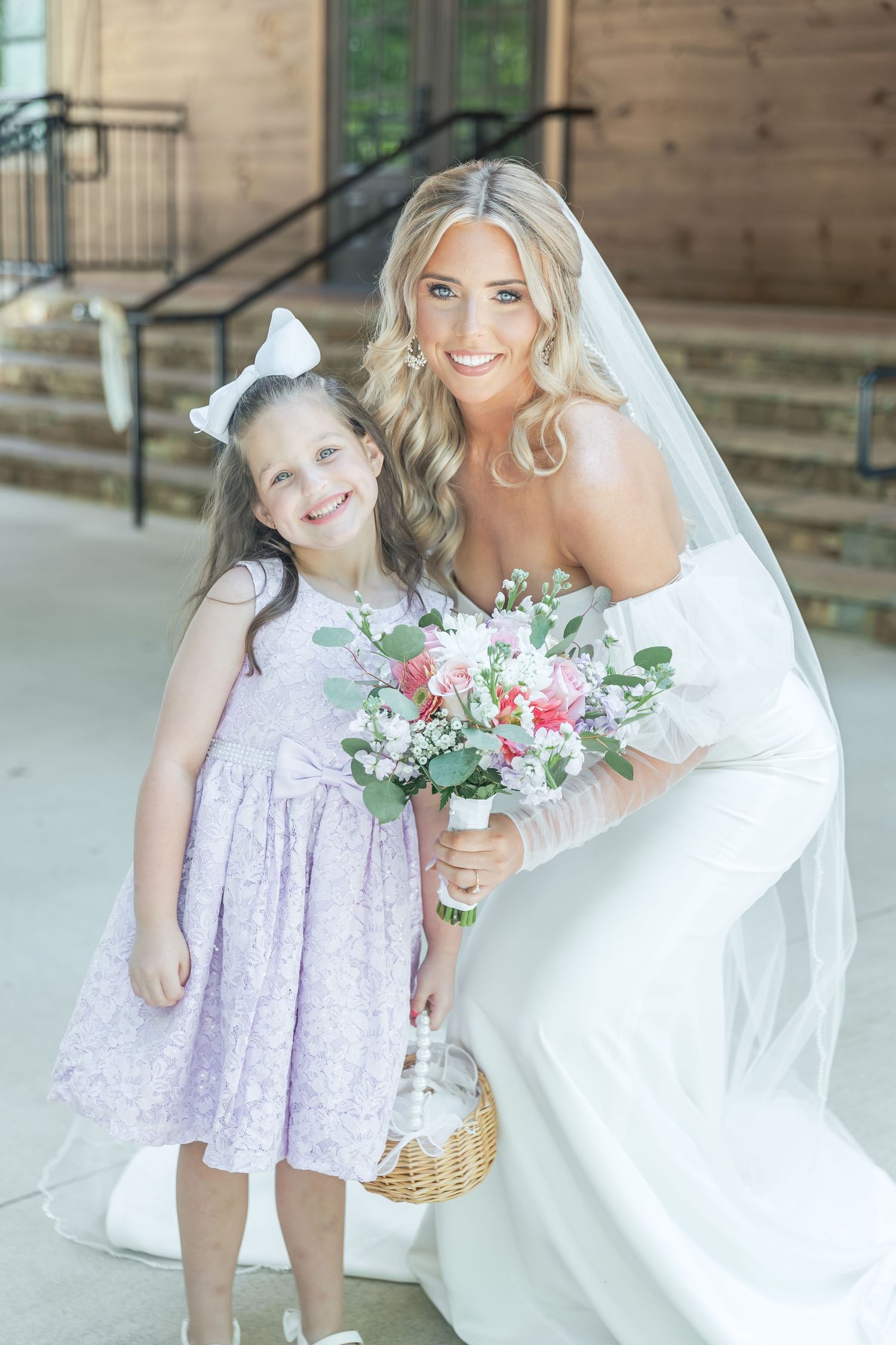 A young flower girl in lavender dress poses with the bride holding a basket on a wooden porch.