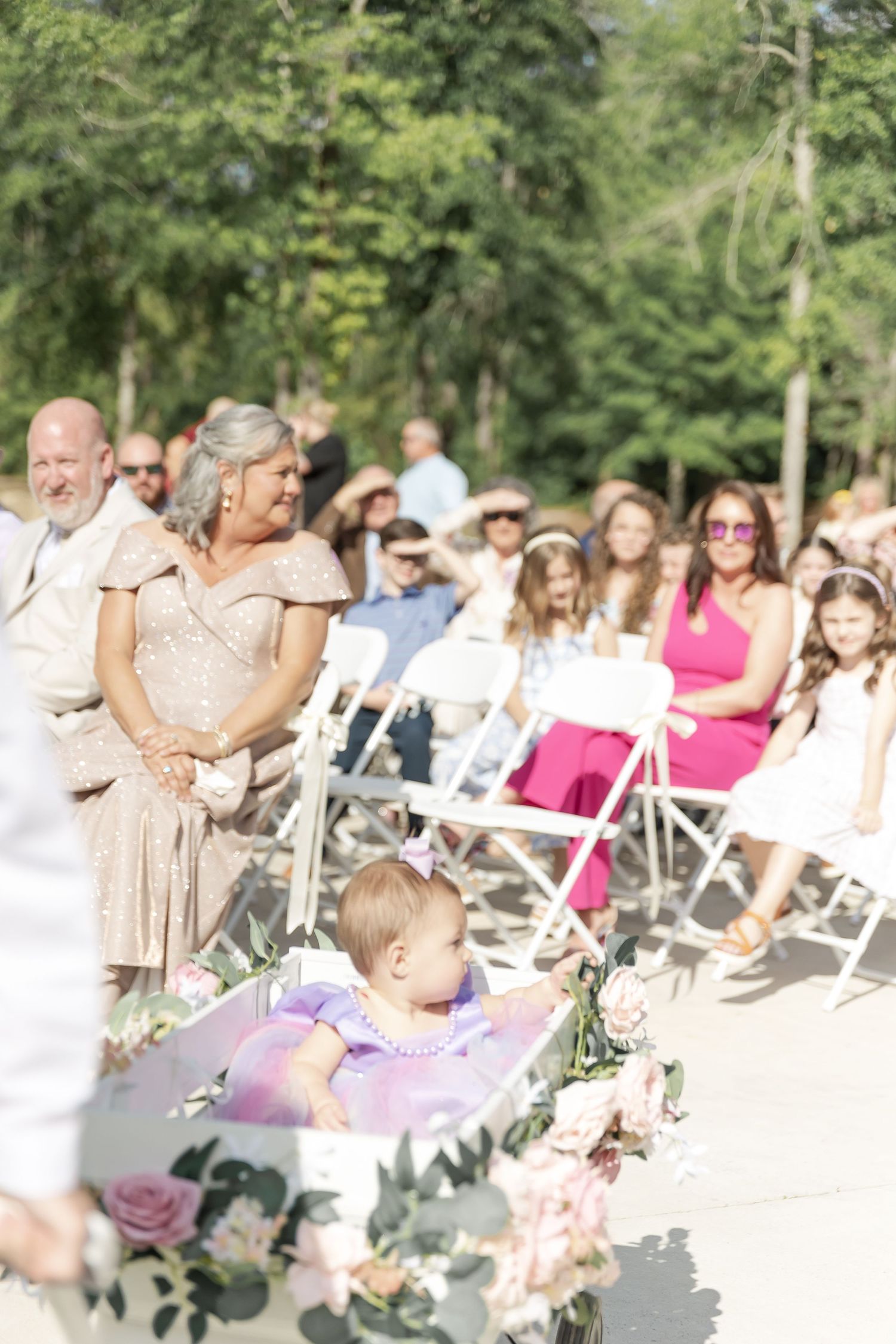 Outdoor wedding ceremony with guests seated in white chairs and floral decorations in a garden setting.