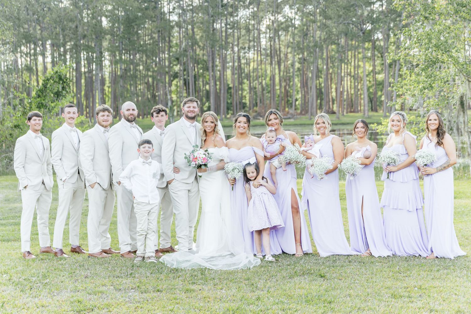 Wedding party in white and lavender attire posed together in front of tall pine trees.