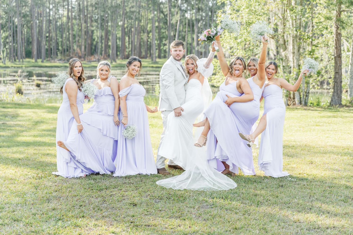 Bridesmaids in flowing lilac dresses pose playfully with couple on grassy lawn.