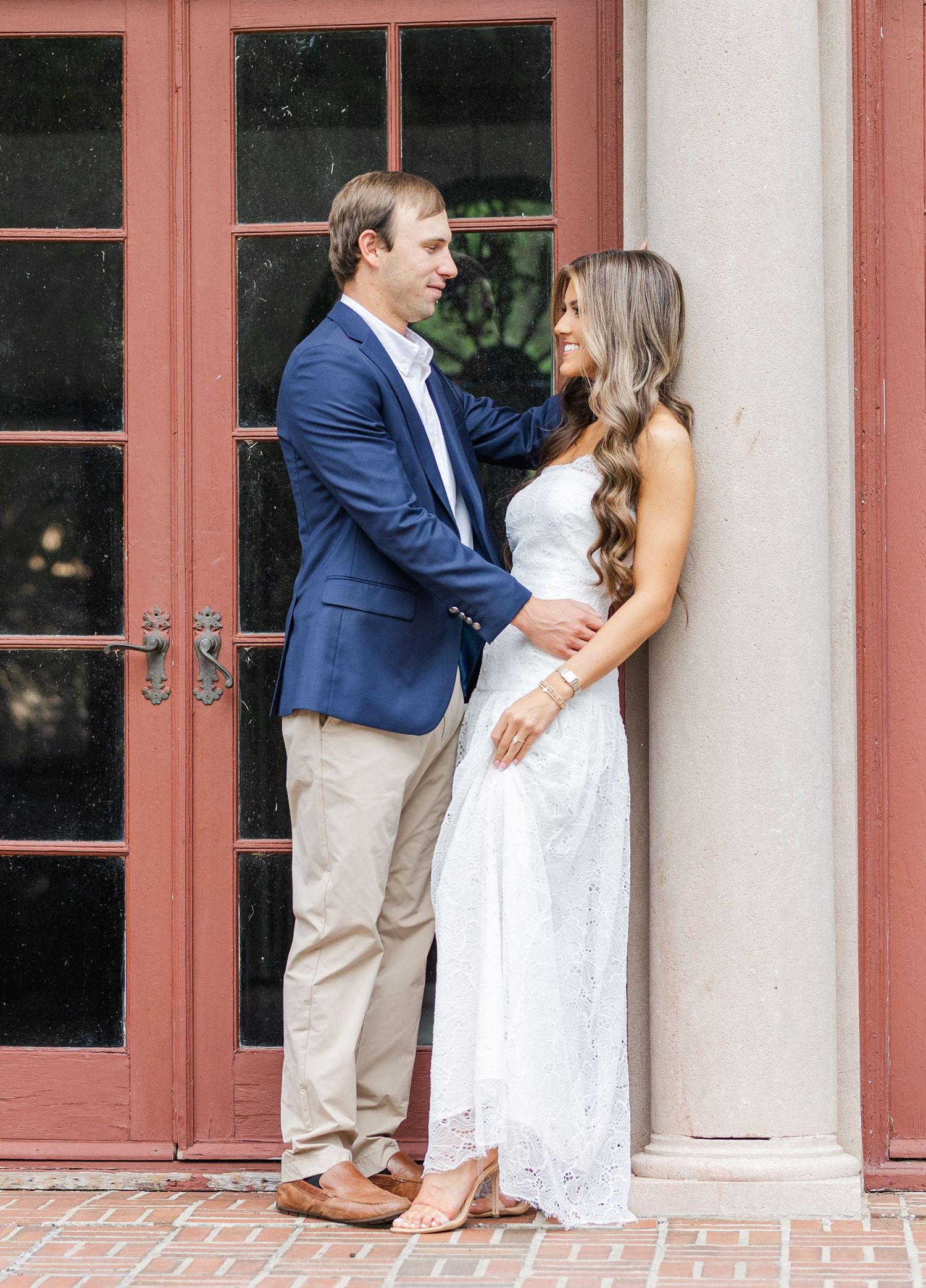Couple in elegant engagement photo, man in navy blazer and woman in white lace dress, posing by red doors.