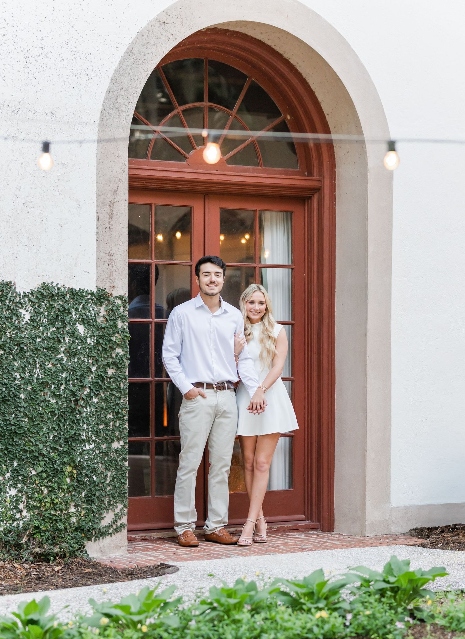 Smiling couple posing together under a grand arched red door entrance with string lights at a historic building.