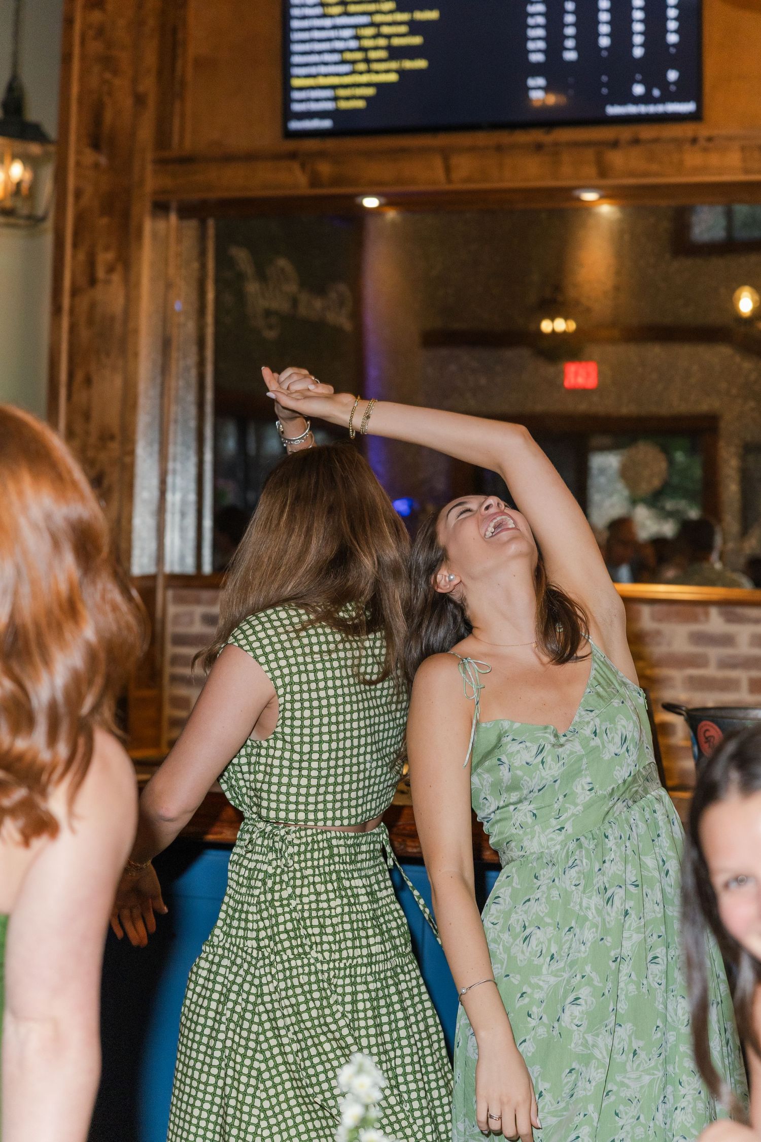 Two women in green dresses dancing joyfully at a bar or restaurant celebration event.