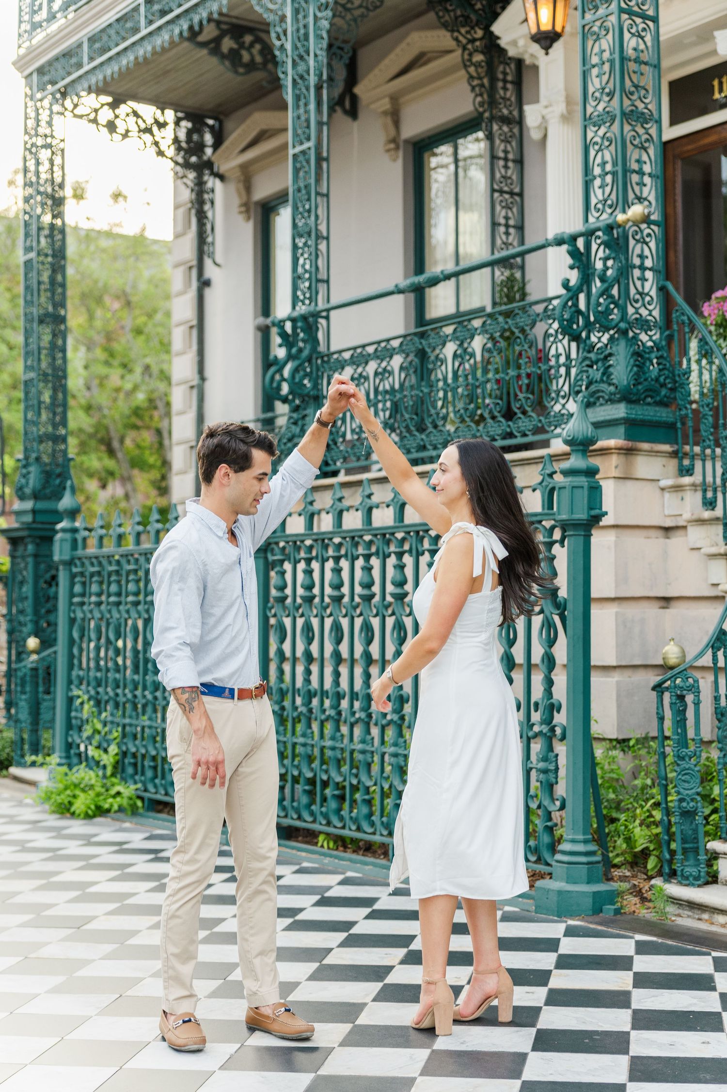 Couple dancing and twirling in front of an ornate green iron fence on a checkered tile porch.