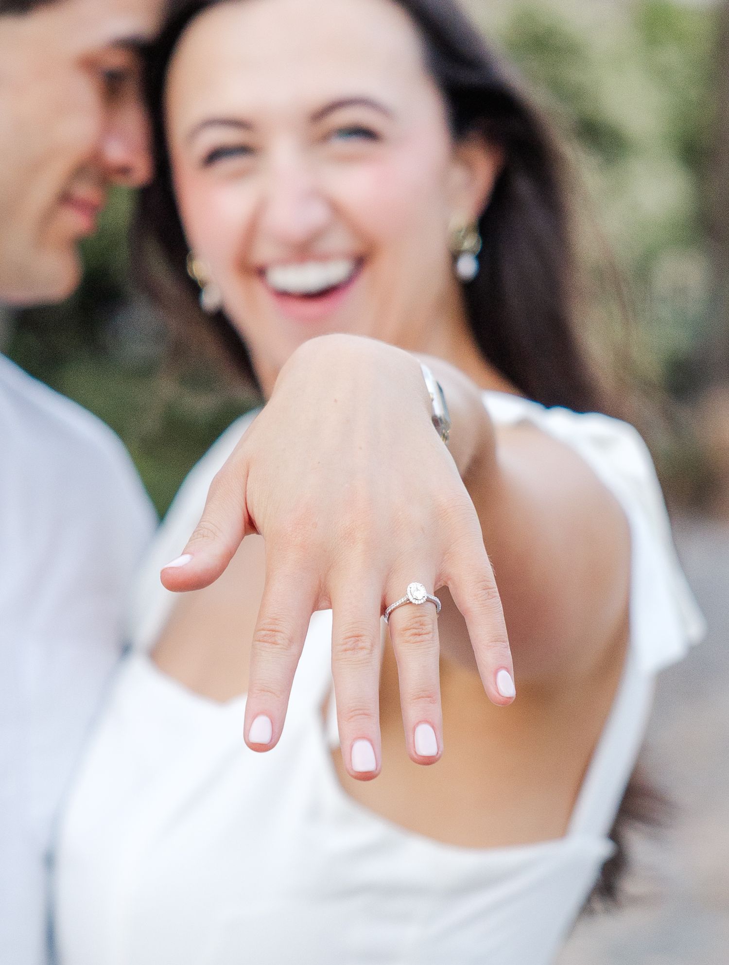 Laughing bride-to-be showing off her diamond engagement ring while embracing her fiance outdoors.