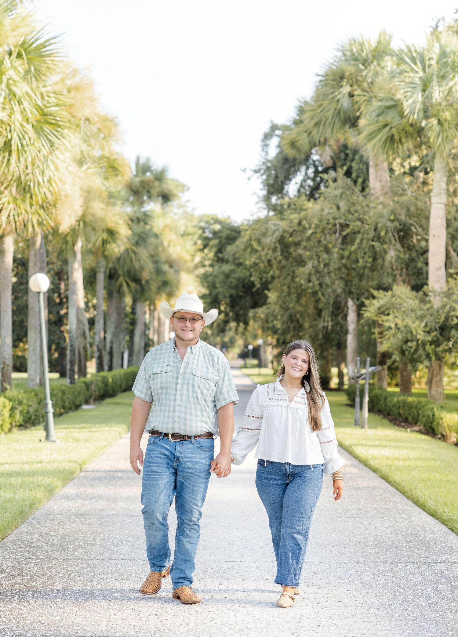 Couple holding hands walking down a palm tree-lined path, man in cowboy hat and woman in white blouse, both wearing jeans.
