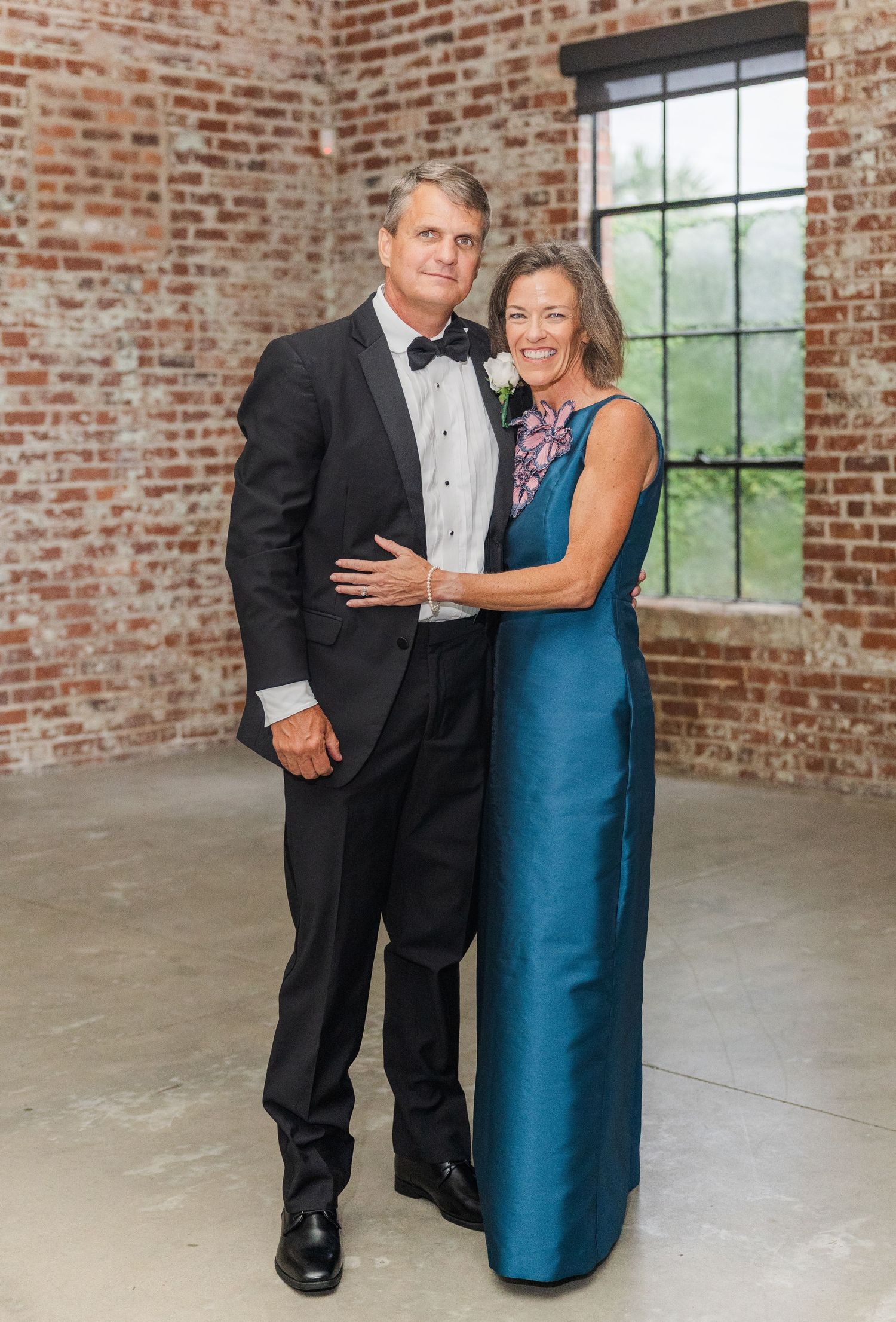 Couple poses together in formal wedding attire against rustic exposed brick wall backdrop.