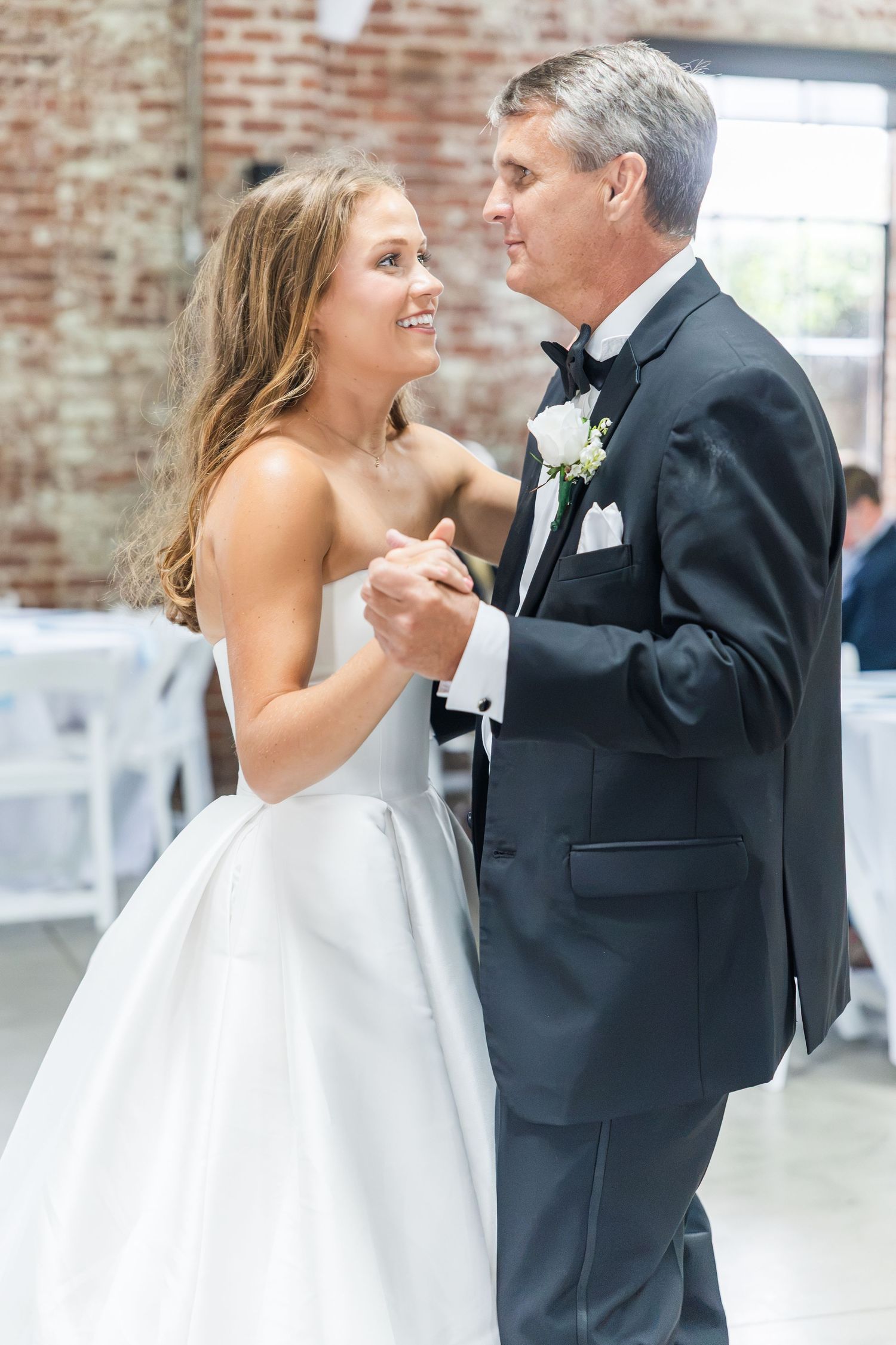 Bride in white ballgown shares father-daughter dance in rustic brick venue during wedding reception.
