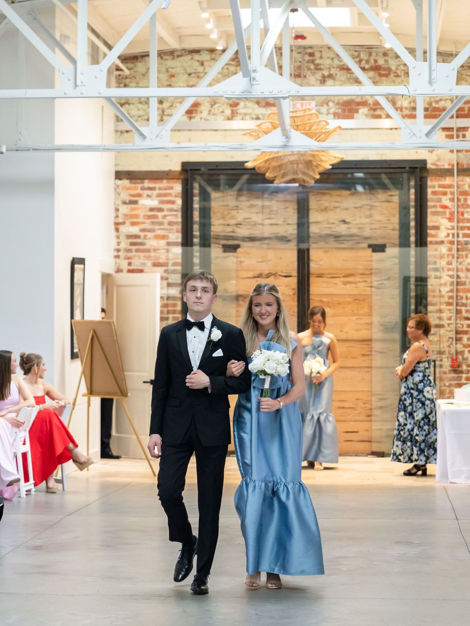 Bridesmaid in blue dress walks down aisle escorted by groomsman in black tuxedo during wedding ceremony.