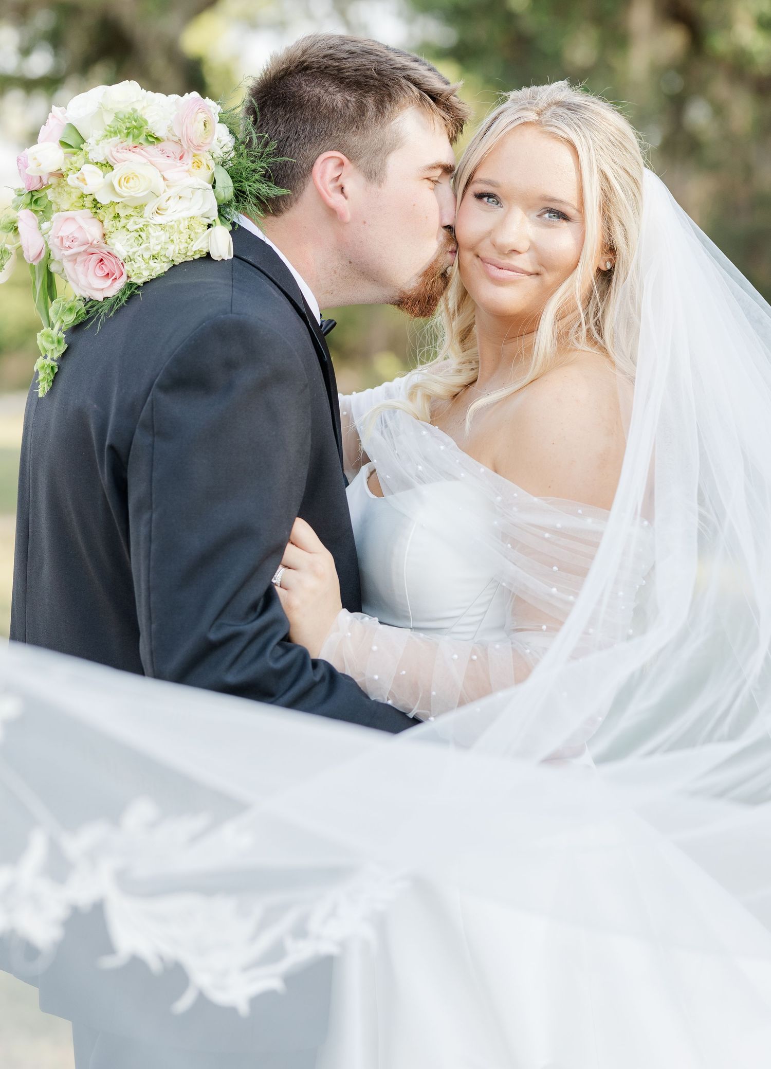 Groom in black tuxedo kissing bride on cheek, bride smiling in lace veil and white gown.