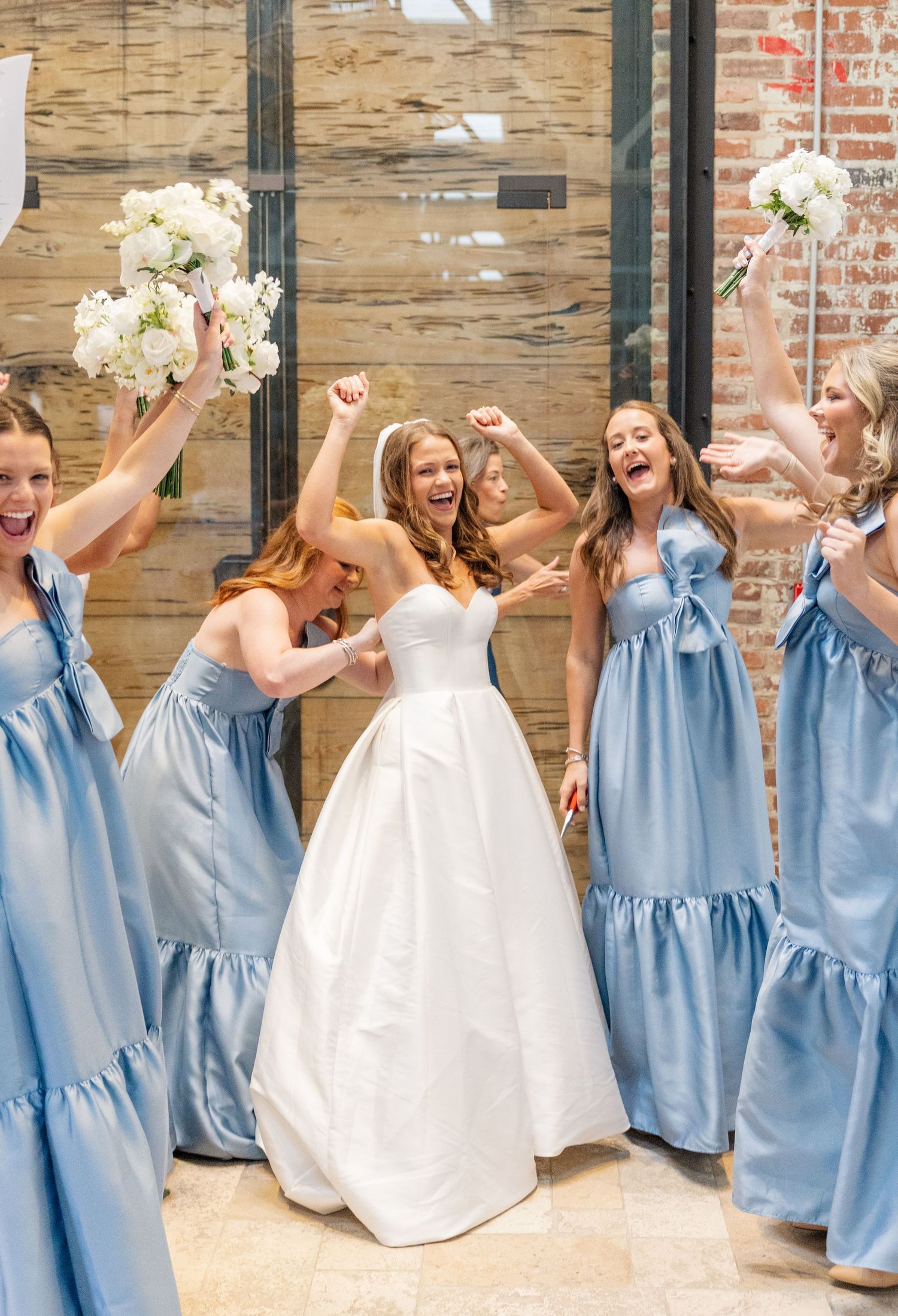 Bride celebrates with bridesmaids in blue dresses raising bouquets joyfully.