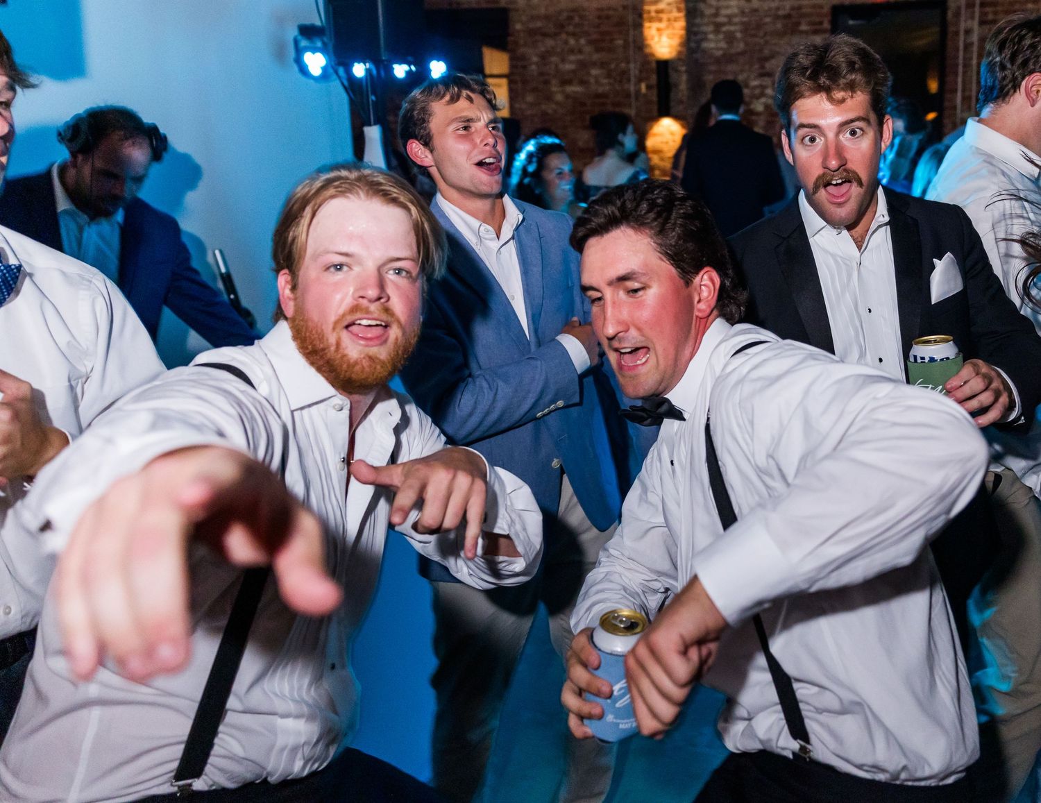 Groomsmen in white shirts and suspenders dance energetically at lively wedding reception.