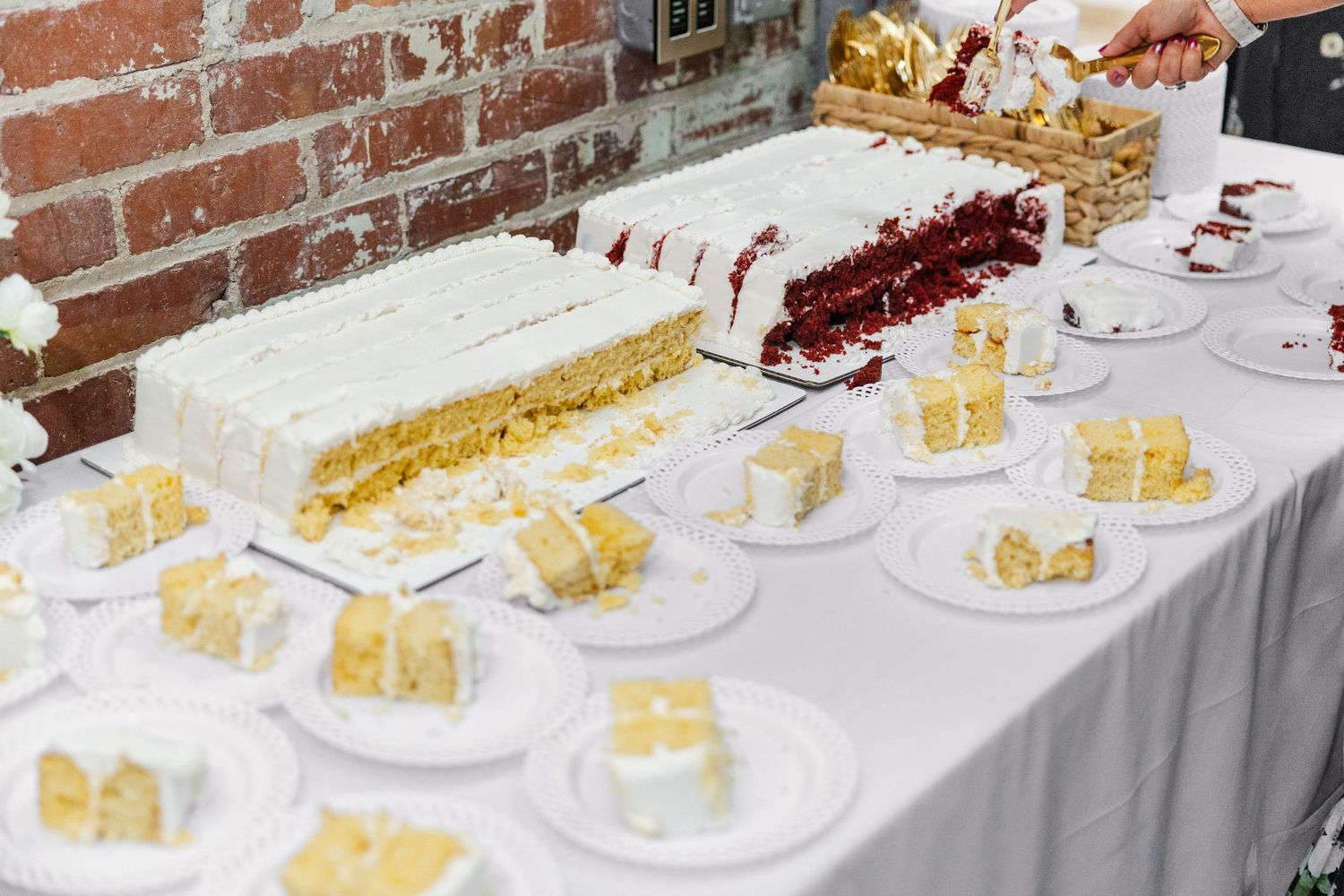 Wedding cake table displays sliced white and yellow sheet cakes on plates against brick wall.