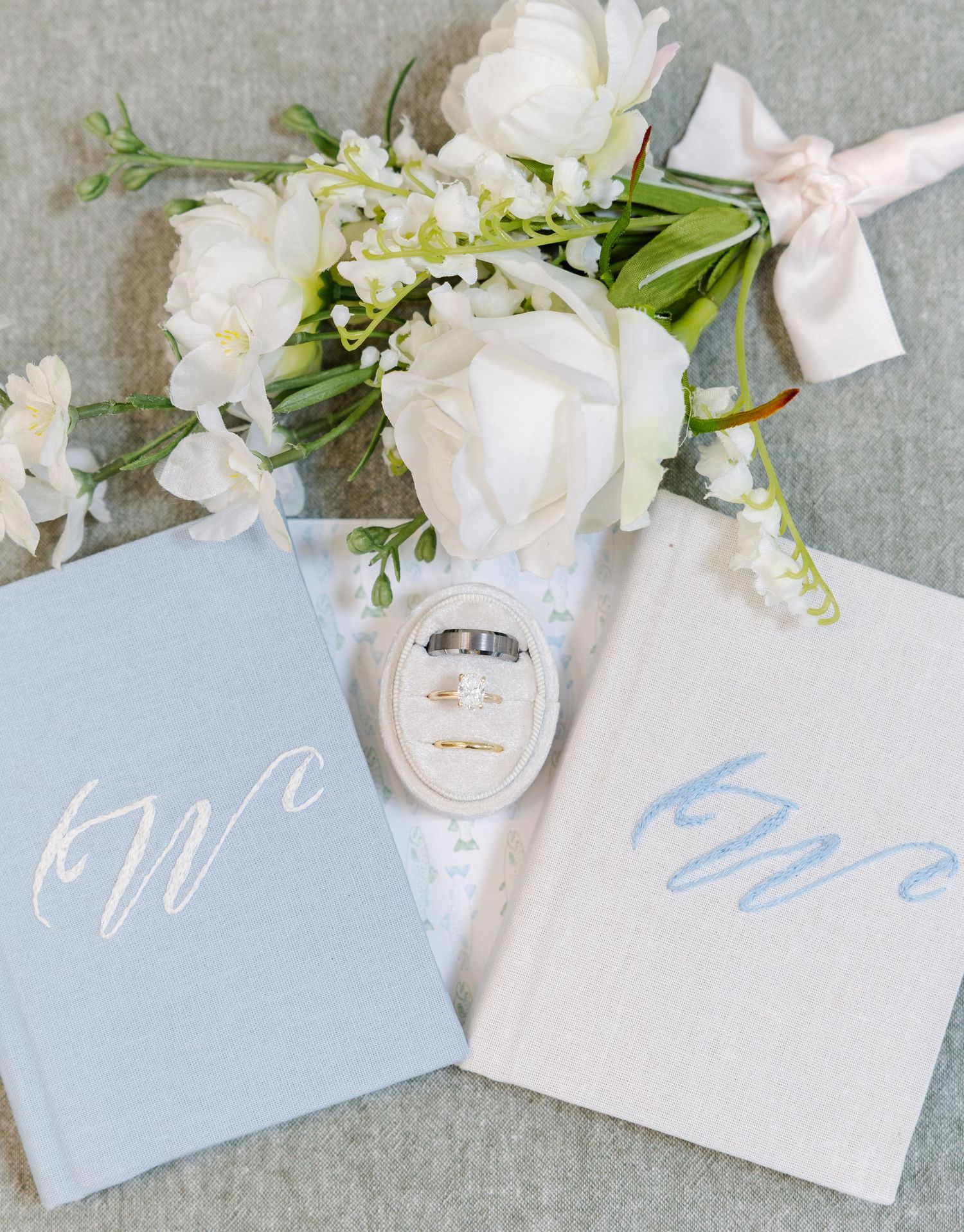 Blue and ivory wedding vow books with calligraphy monograms, gold rings in velvet box, and white floral bouquet.