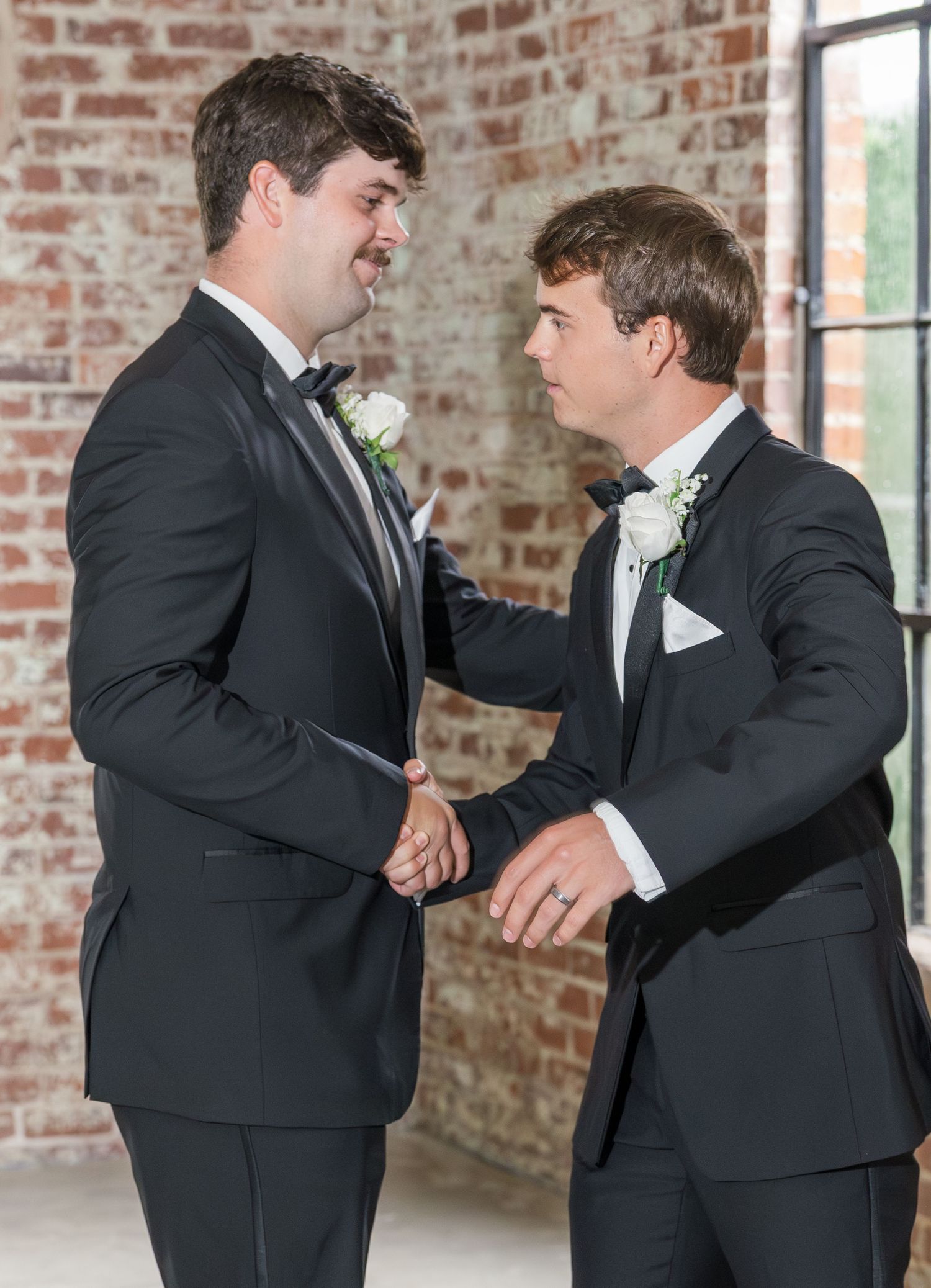 Two groomsmen in black tuxedos sharing a moment together against an exposed brick wall.