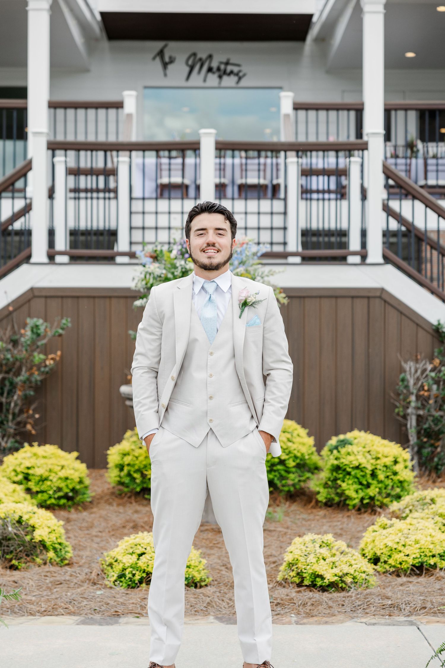 Dapper groom in tan three-piece suit with light blue tie poses confidently outside wedding venue.
