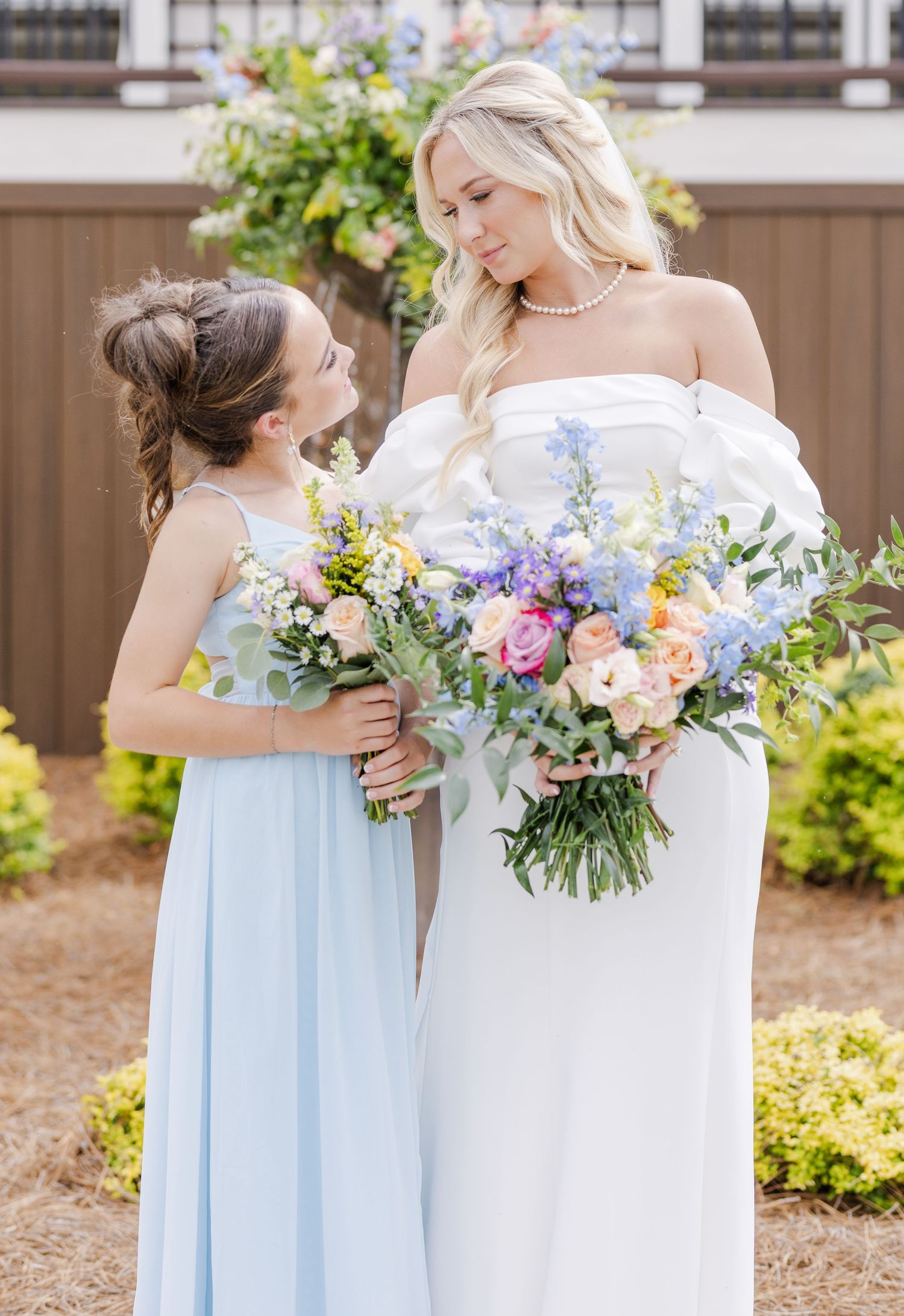 Blonde bride in white strapless gown and young girl in light blue dress holding colorful bouquets.
