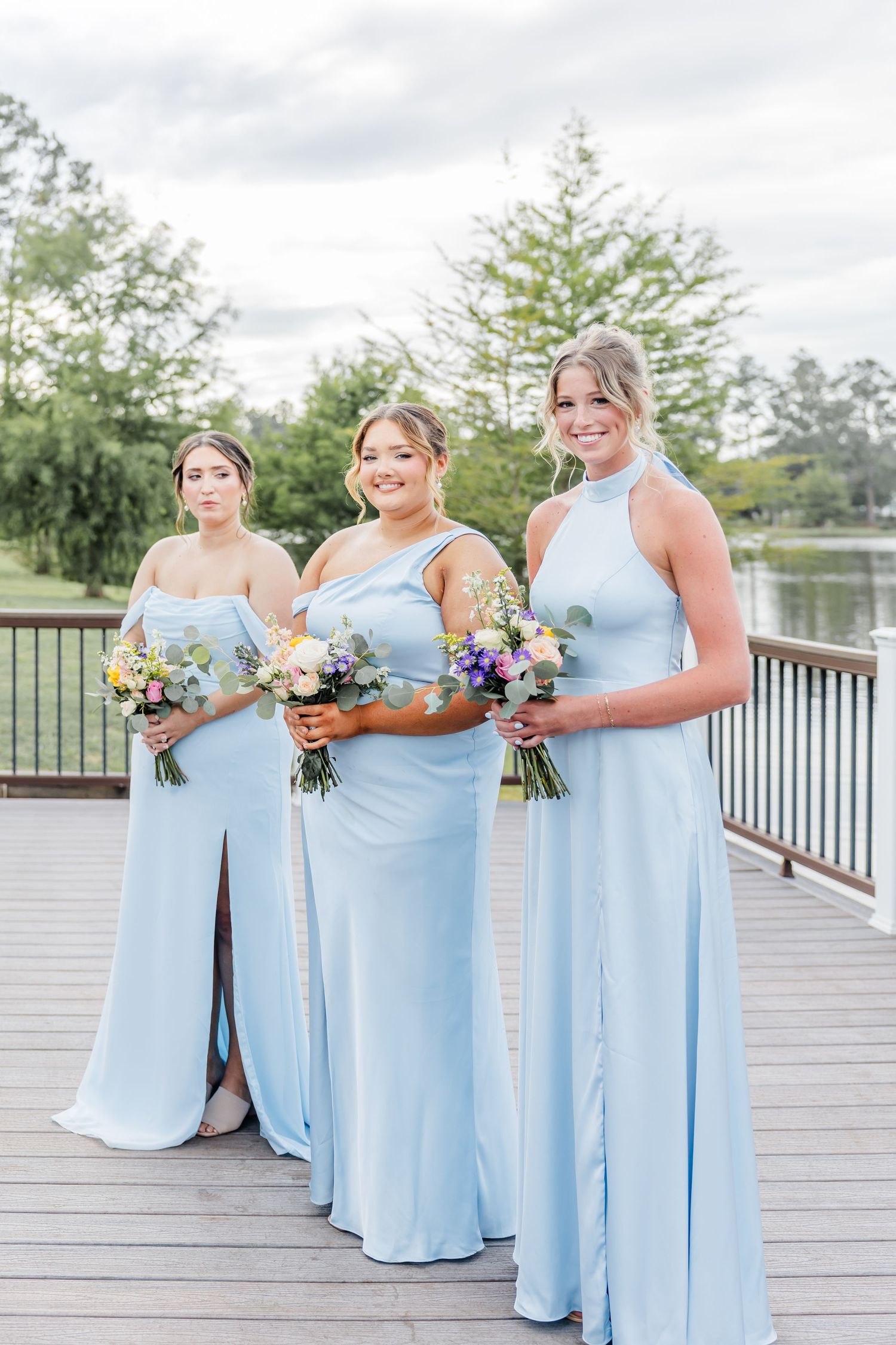 Three bridesmaids in light blue gowns holding colorful bouquets on lakeside wooden deck.