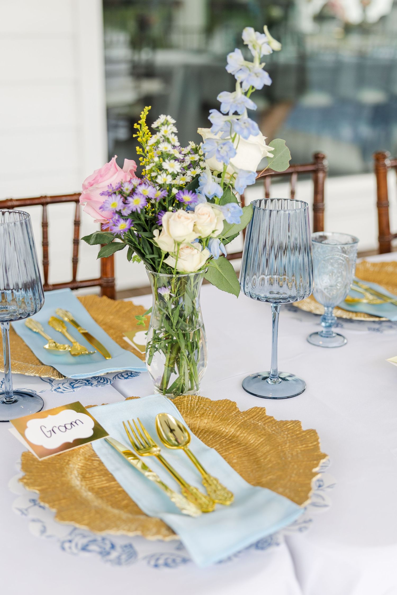Elegant wedding table setting with gold charger plates, blue glassware, gold flatware, and a floral centerpiece with blue and white blooms.