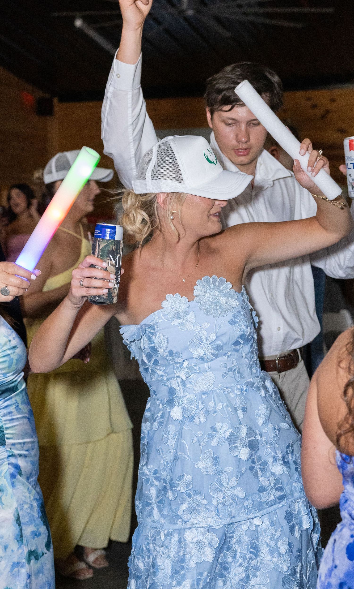 Bride in a blue floral dress dances with guests holding glow sticks at a lively wedding reception party.