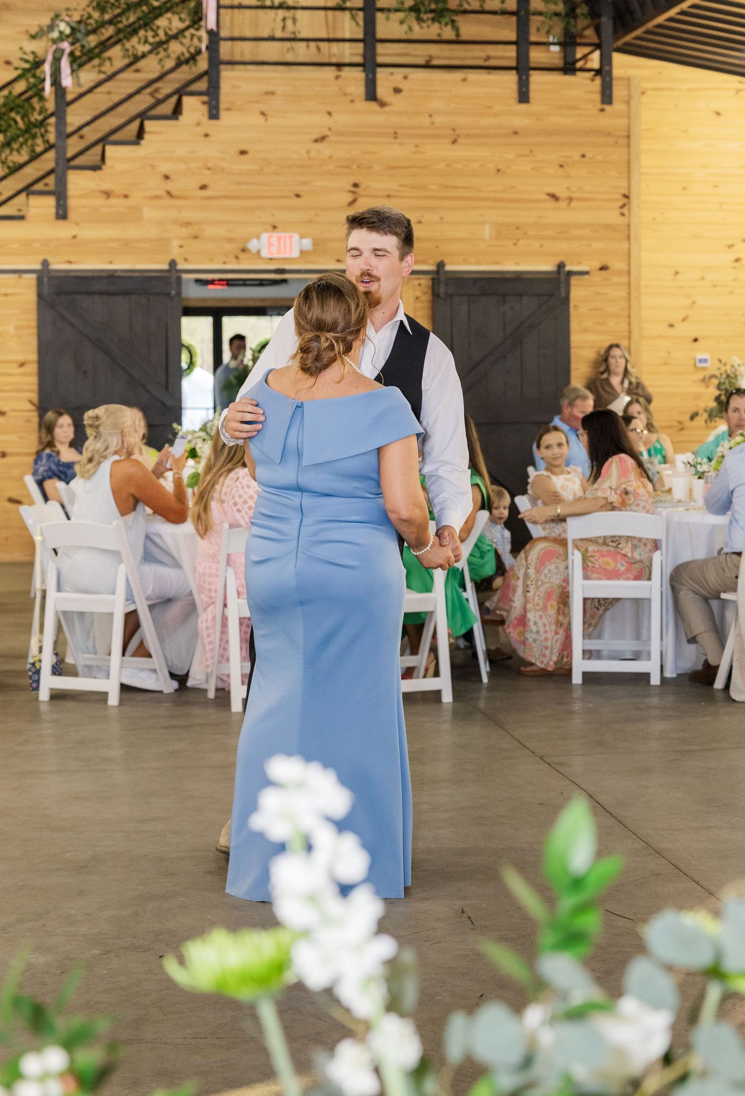 Groom dances with woman in blue gown during mother-son dance at rustic barn wedding reception.