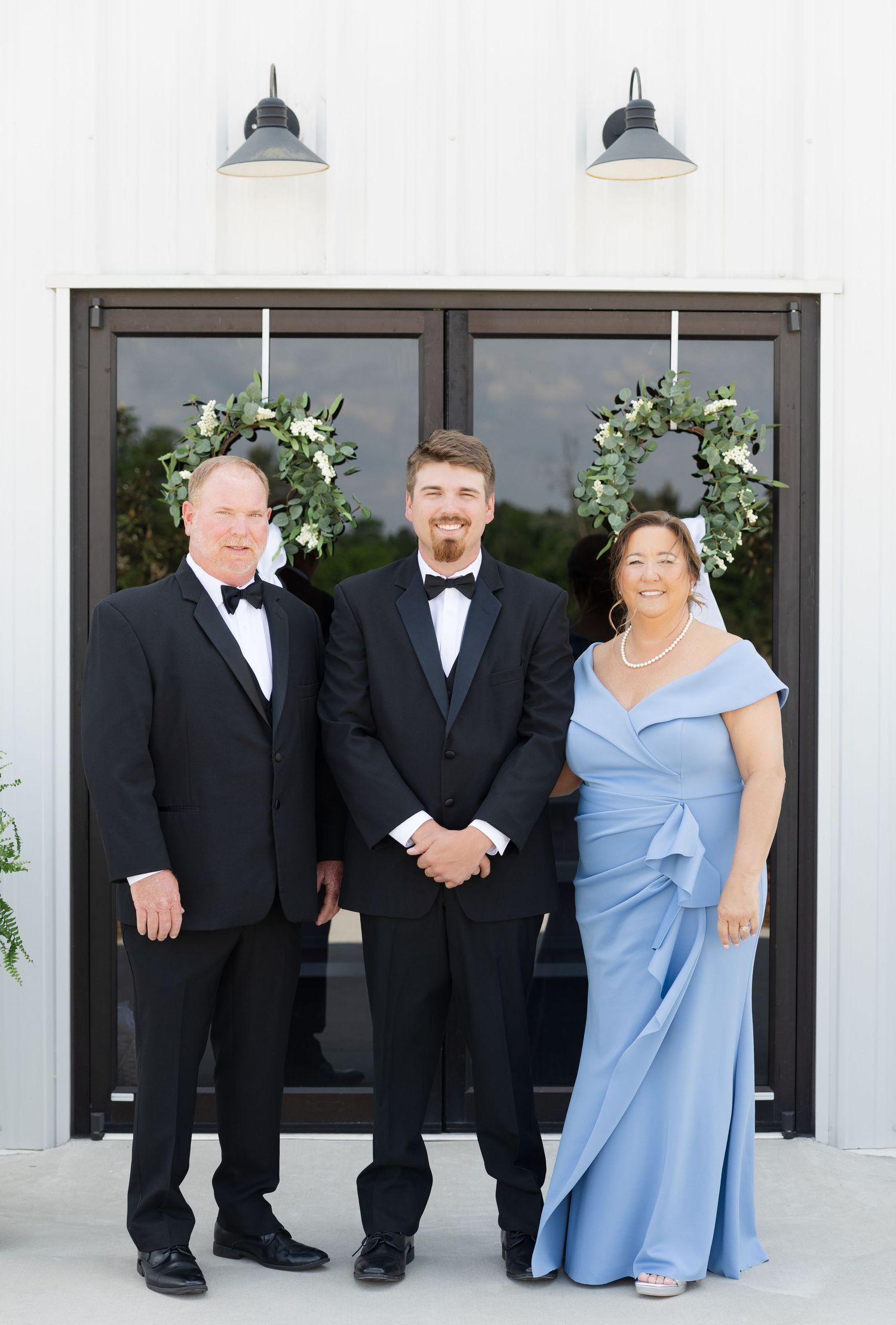 Groom in tuxedo poses with parents in formal attire outside white barn wedding venue.