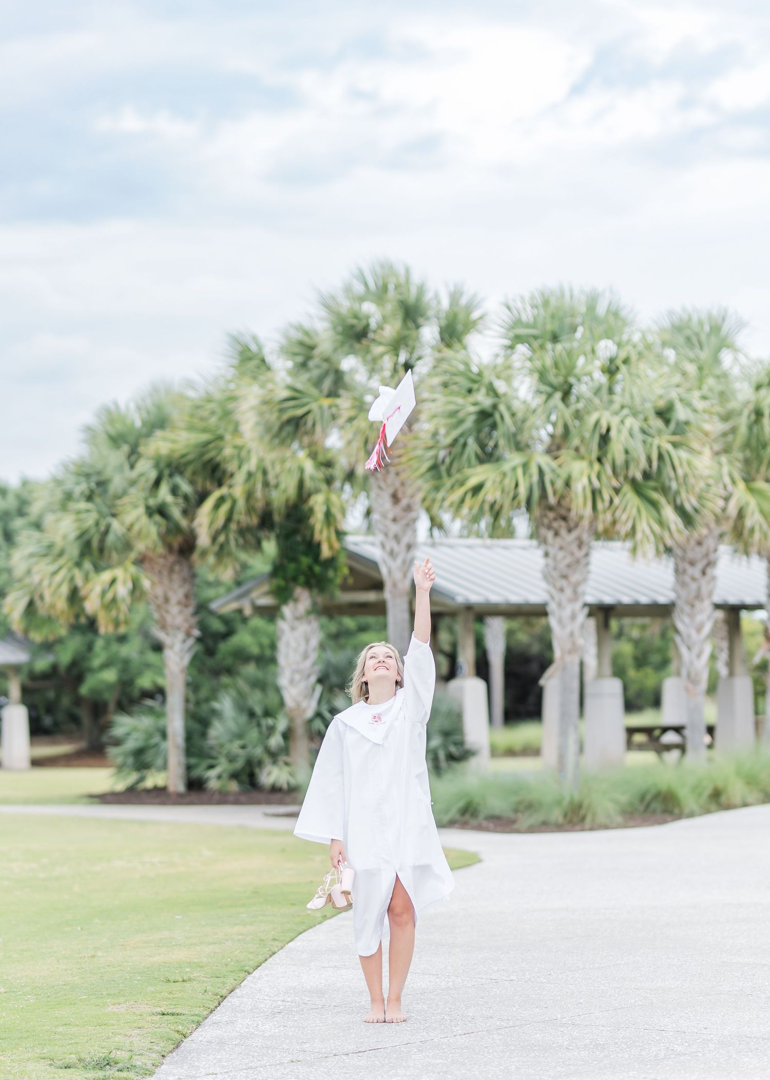 Graduate in white dress poses playfully on a path surrounded by tall palm trees.
