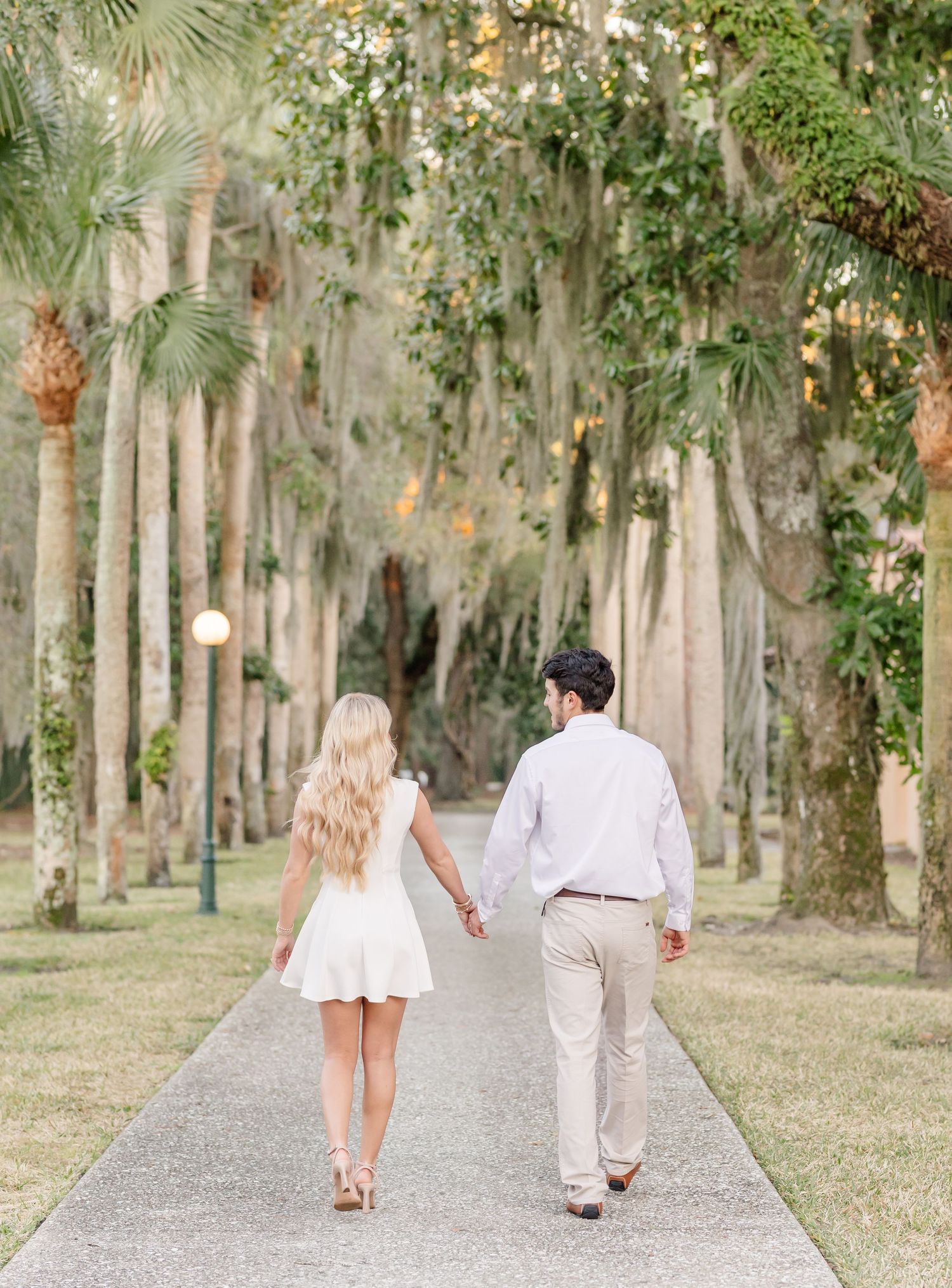 A couple in white clothing walks hand in hand down a palm tree-lined path at sunset.