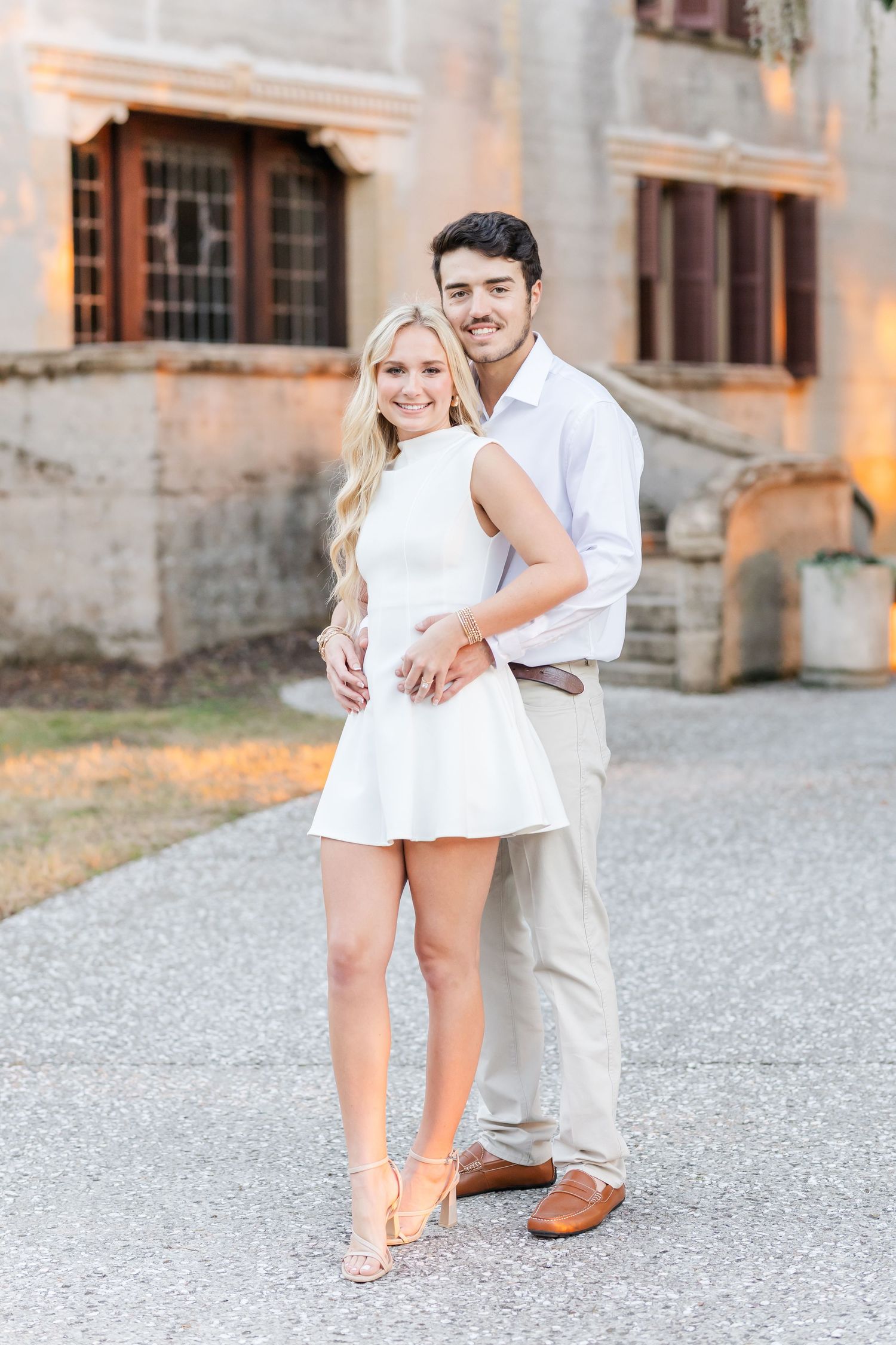 A couple in coordinated white and beige outfits pose together on a gravel path in front of a rustic building.