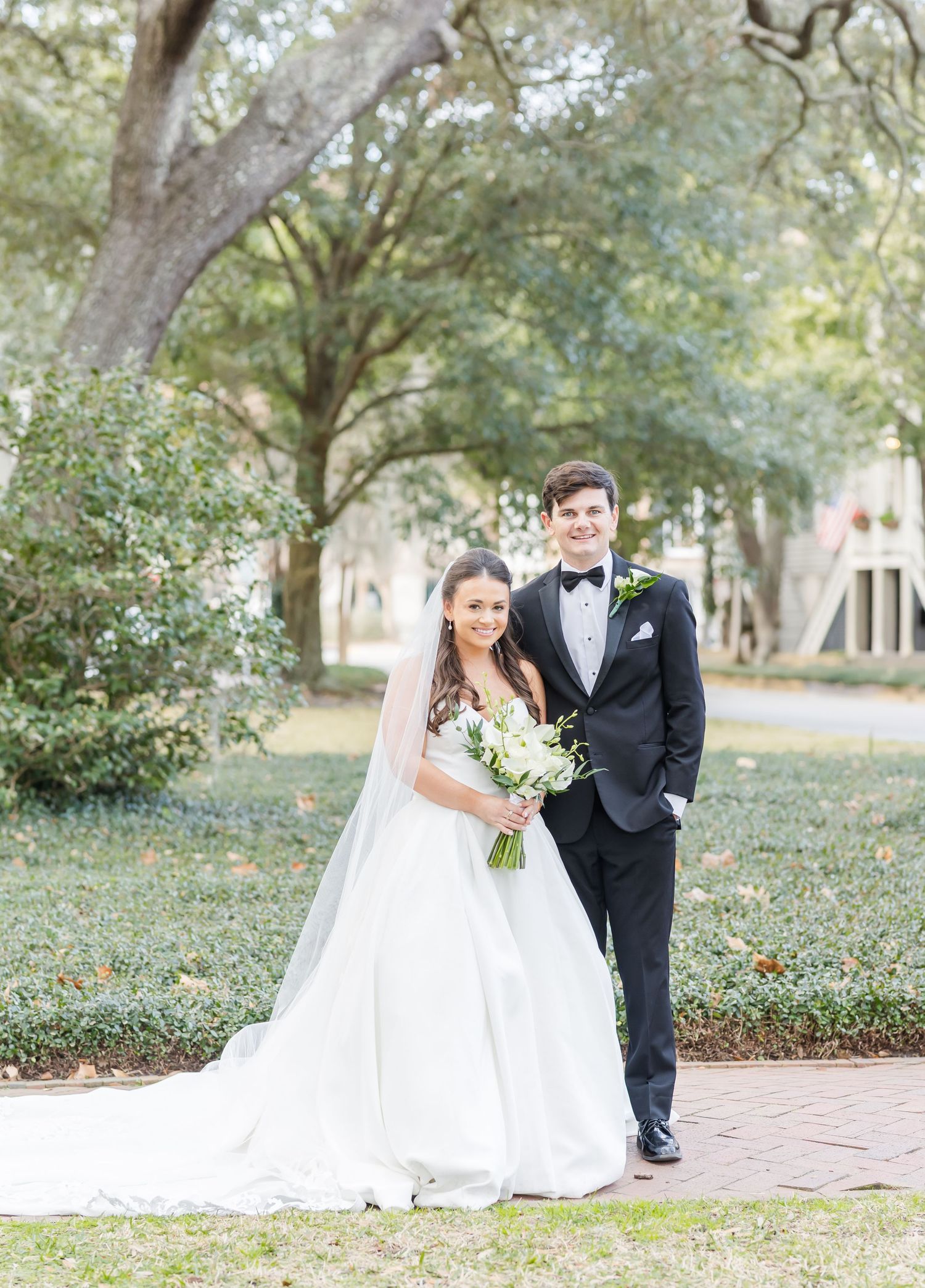 A wedding couple poses together in a scenic outdoor garden setting with trees and brick pathways.