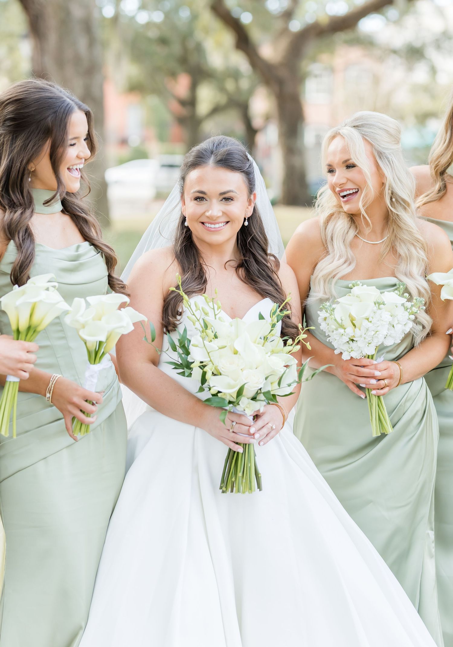 Bridesmaids in sage green dresses hold white bouquets while posing with bride in white gown at outdoor wedding.