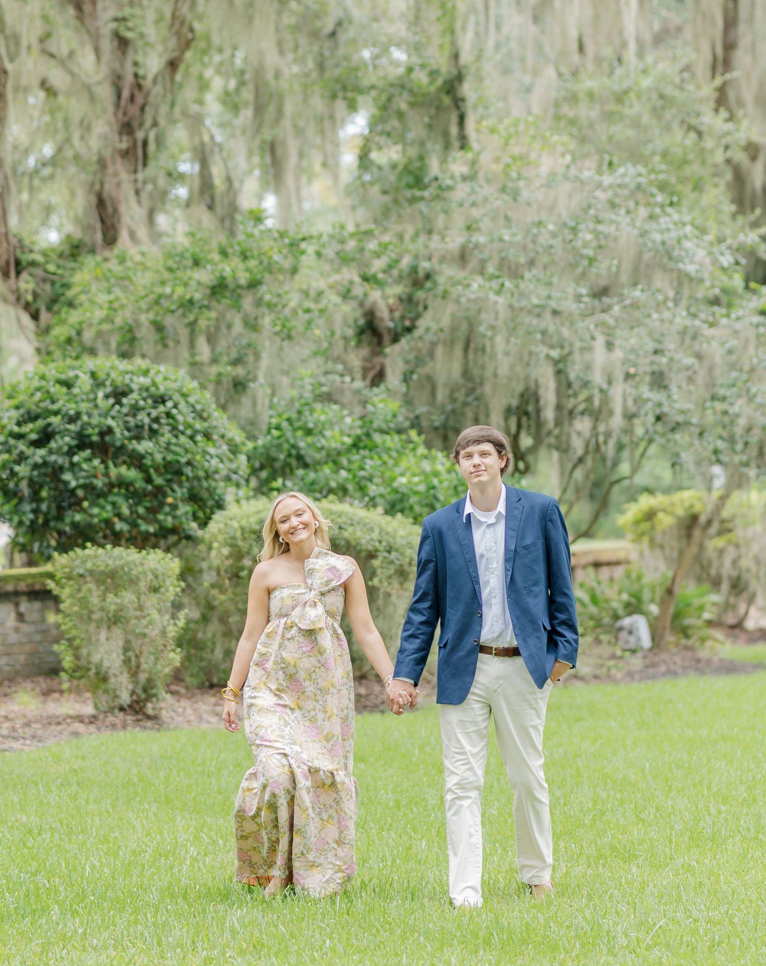 Couple in spring attire walking and posing together on a grassy lawn surrounded by moss-draped trees.