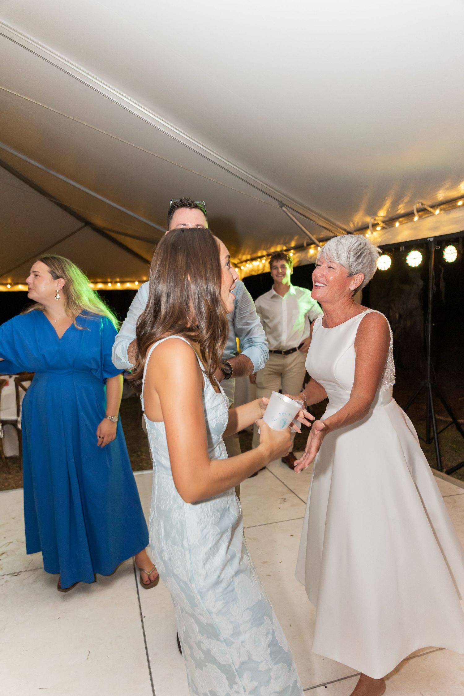 People dancing and mingling at a wedding celebration under a white tent with ambient lighting.