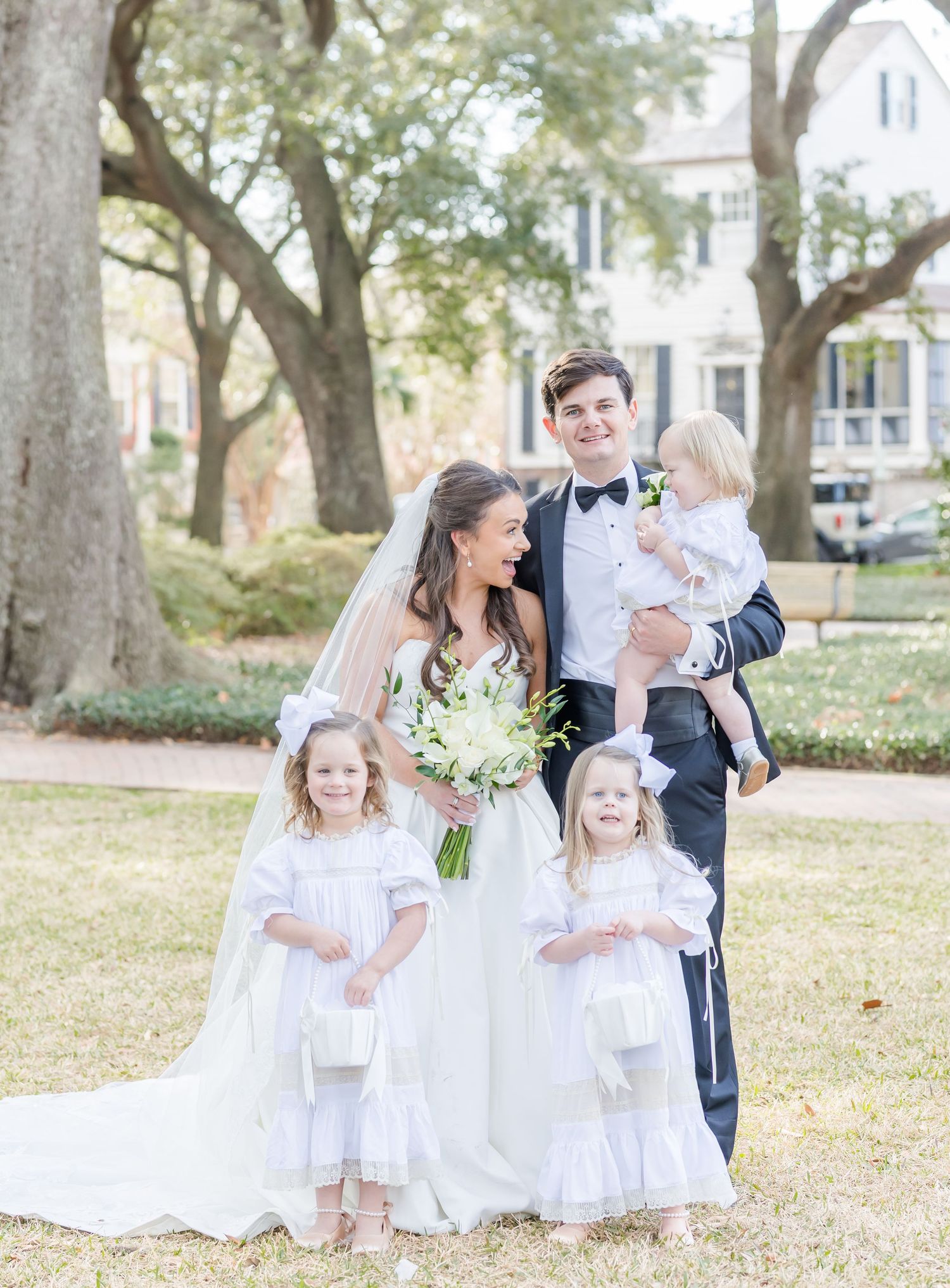 Young flower girls in white dresses pose with the wedding couple on a grassy lawn.