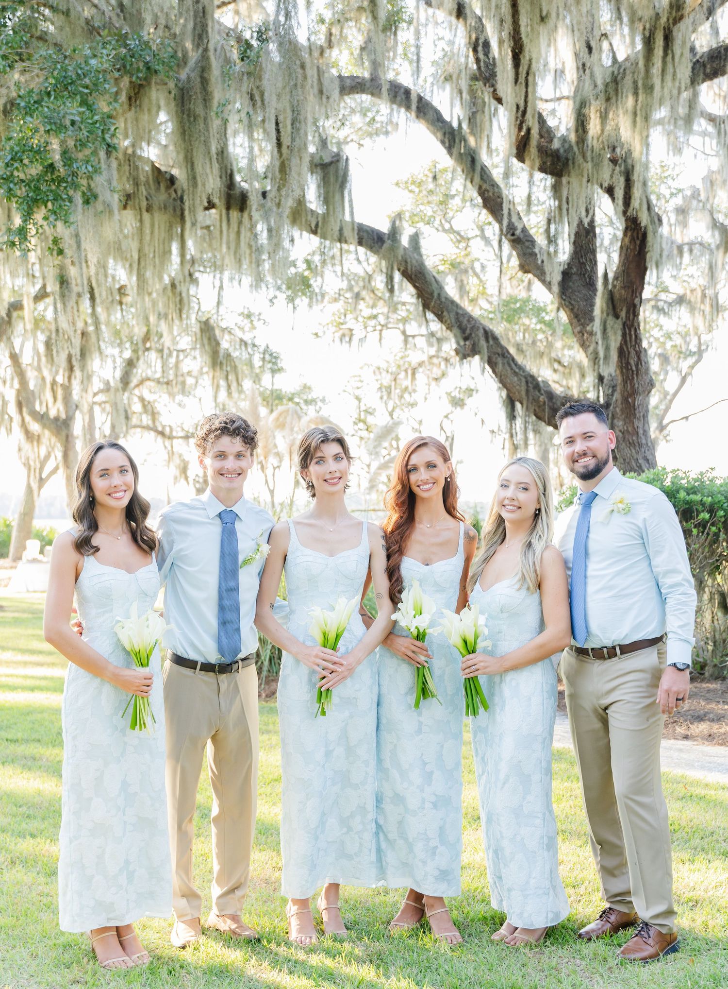 Wedding party in white lace dresses and light suits poses under Spanish moss draped trees at outdoor venue.