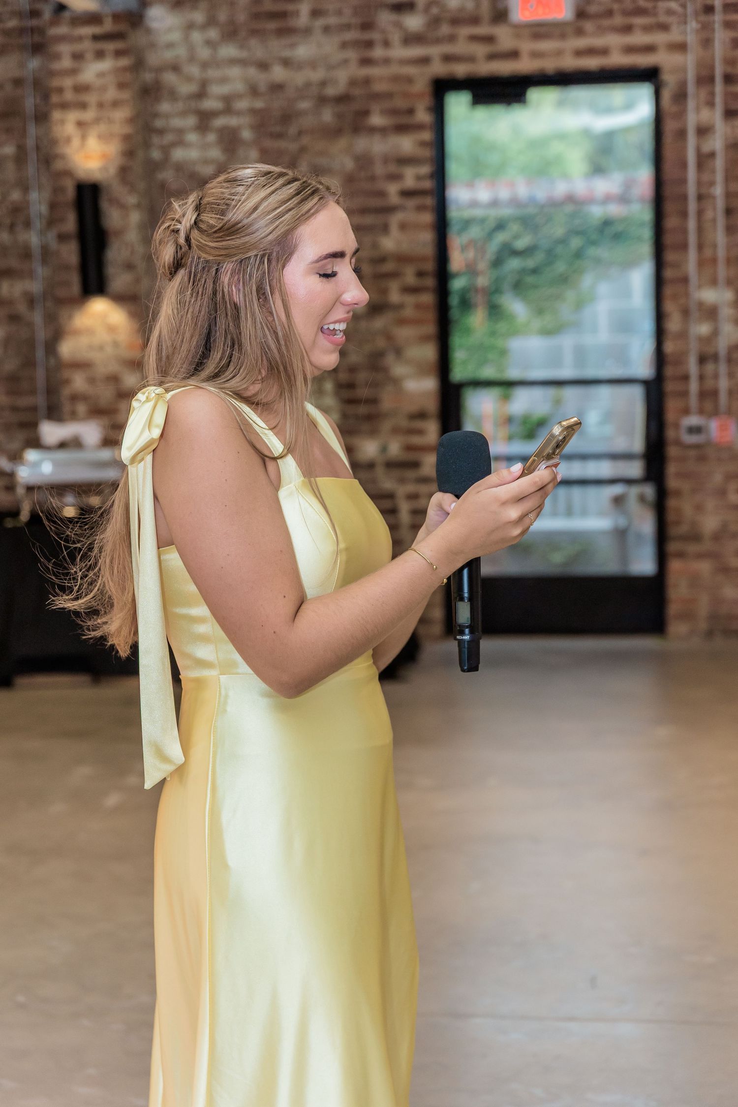 Person in yellow dress holding microphone and phone in rustic brick venue.