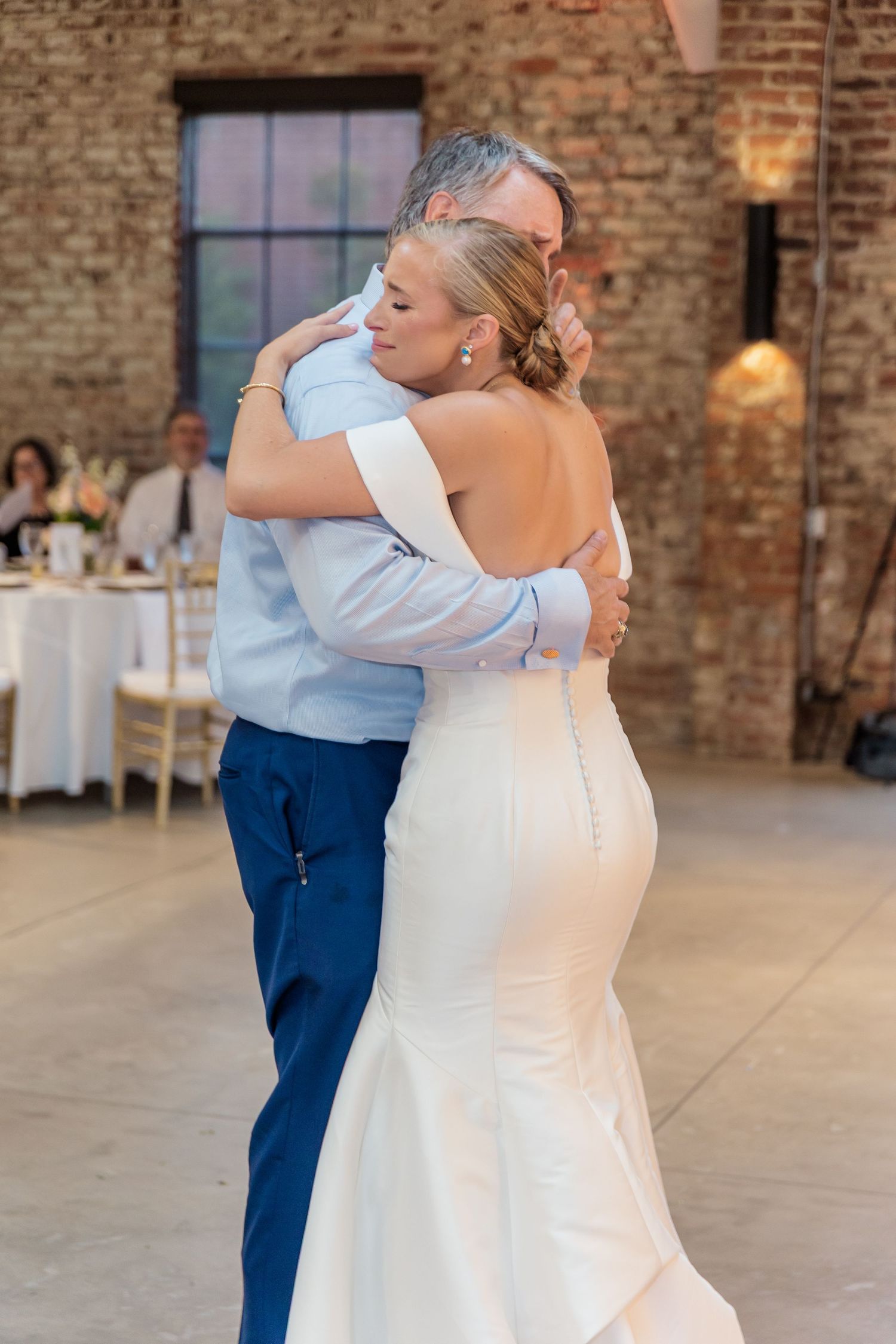 A couple shares a dance in a rustic brick-walled venue with white tables in the background.