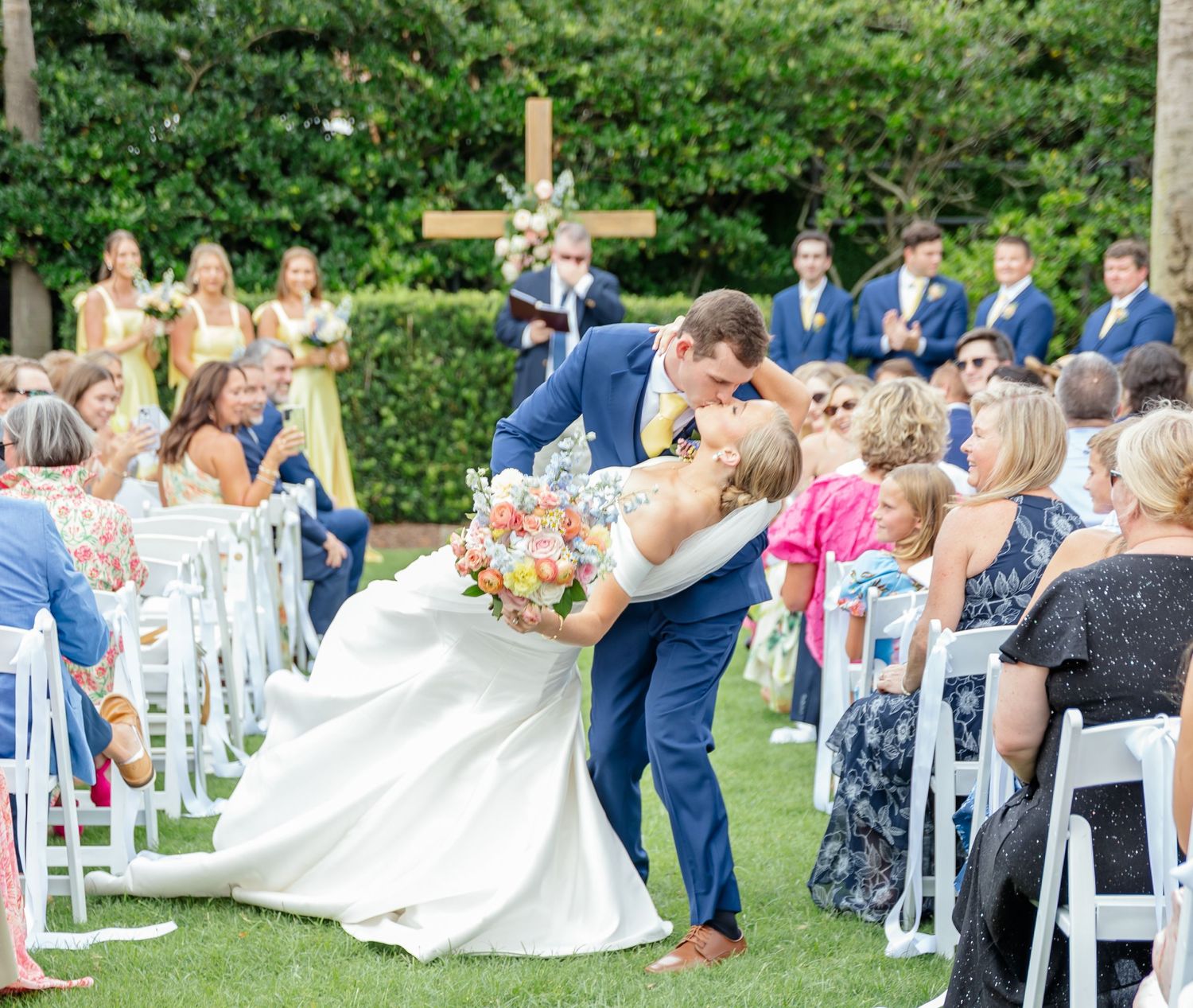 A couple shares their first kiss as newlyweds during an outdoor garden wedding ceremony with guests and wedding party looking on.