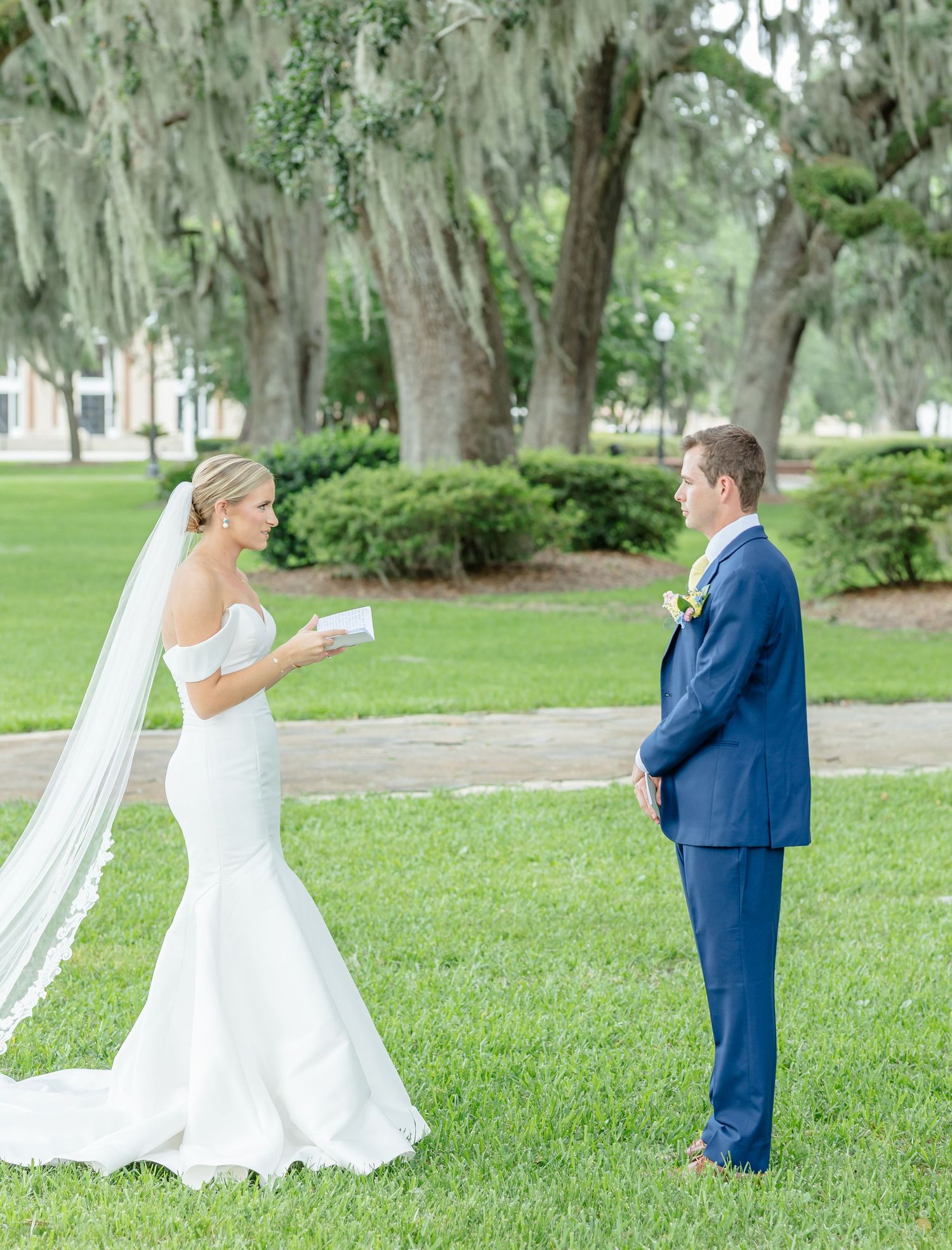 A couple in wedding attire playfully interact on a grassy lawn surrounded by Spanish moss-draped trees.