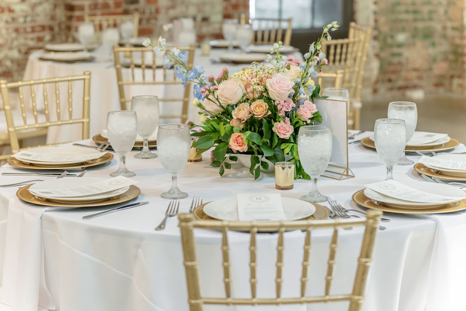 Elegant wedding reception table settings with gold chargers, white linens, and pink floral centerpieces in a rustic venue.