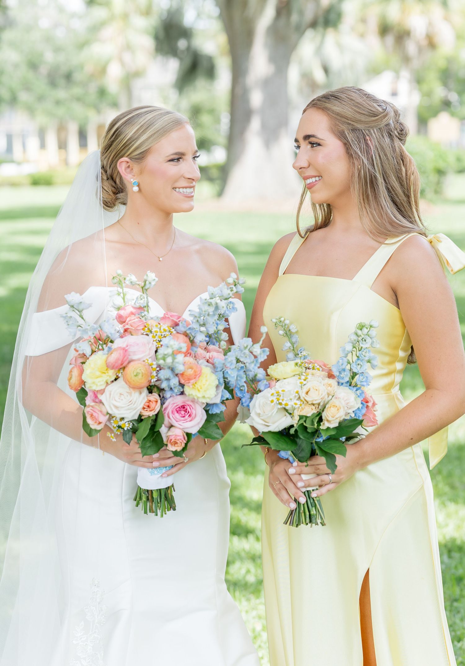A bride in white and bridesmaid in yellow holding vibrant pastel bouquets with roses and hydrangeas in a garden setting.