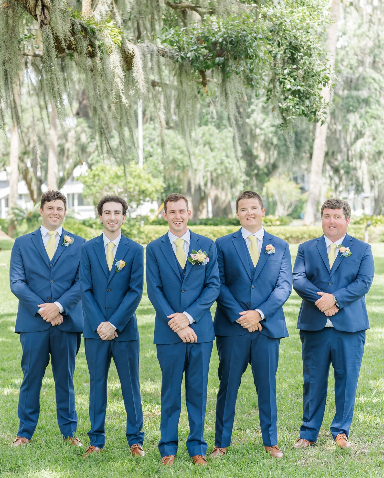 Groomsmen in matching navy blue suits and yellow ties show off colorful socks while posing on grass under Spanish moss trees.