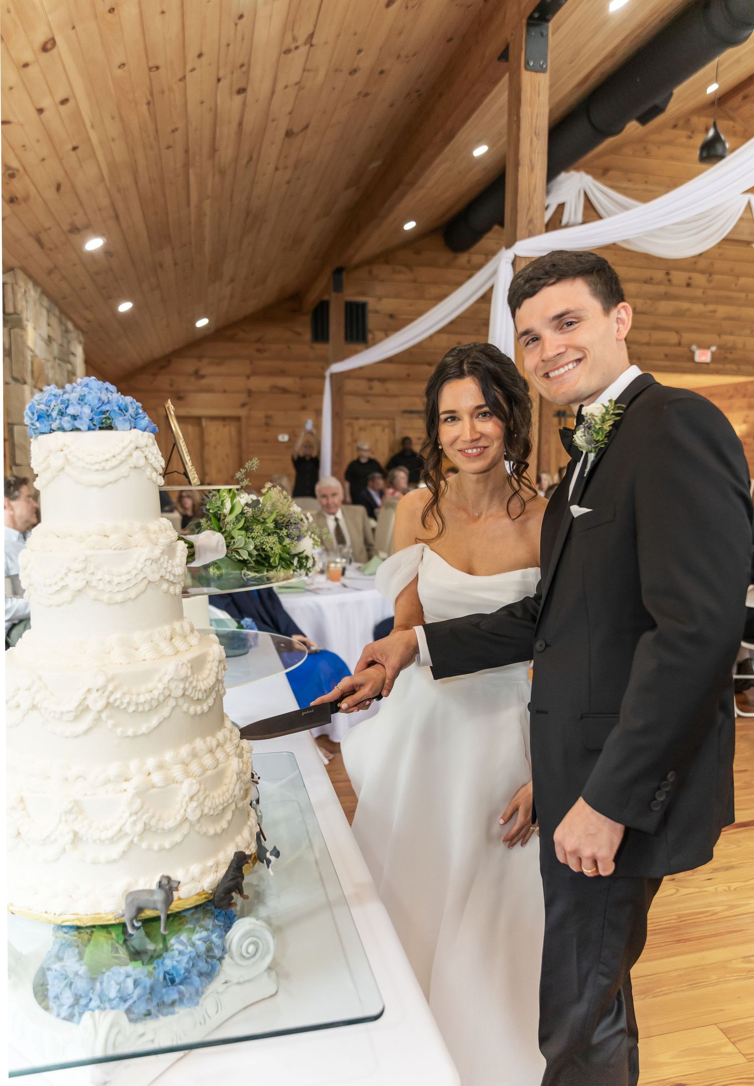 A couple cuts their elegant white wedding cake decorated with blue flowers in a rustic venue.