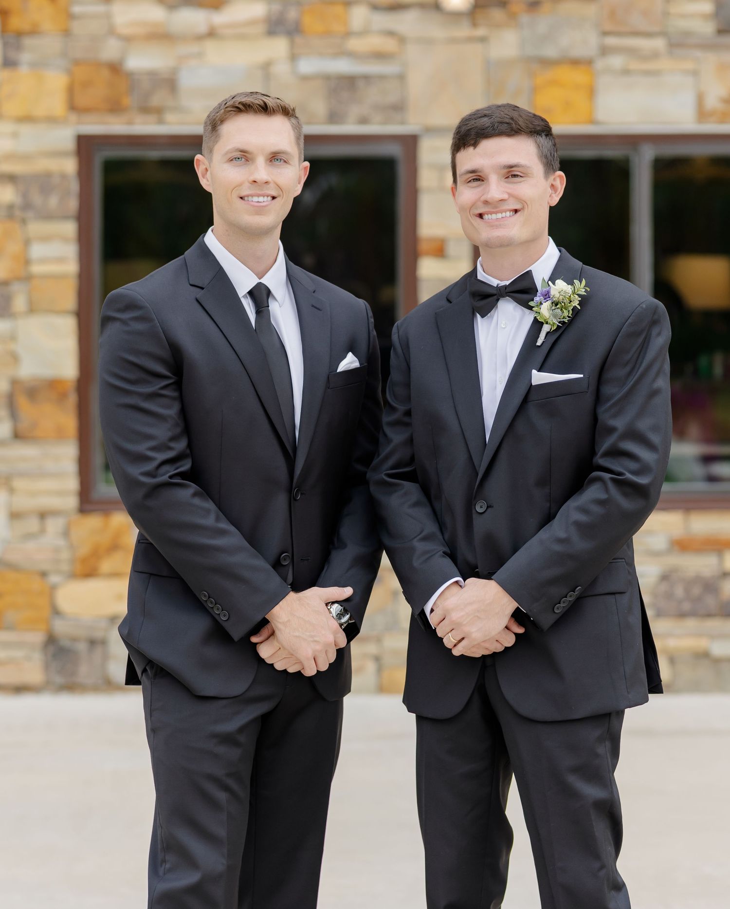 Groomsmen share laughs while posing in matching black tuxedos outside stone building.