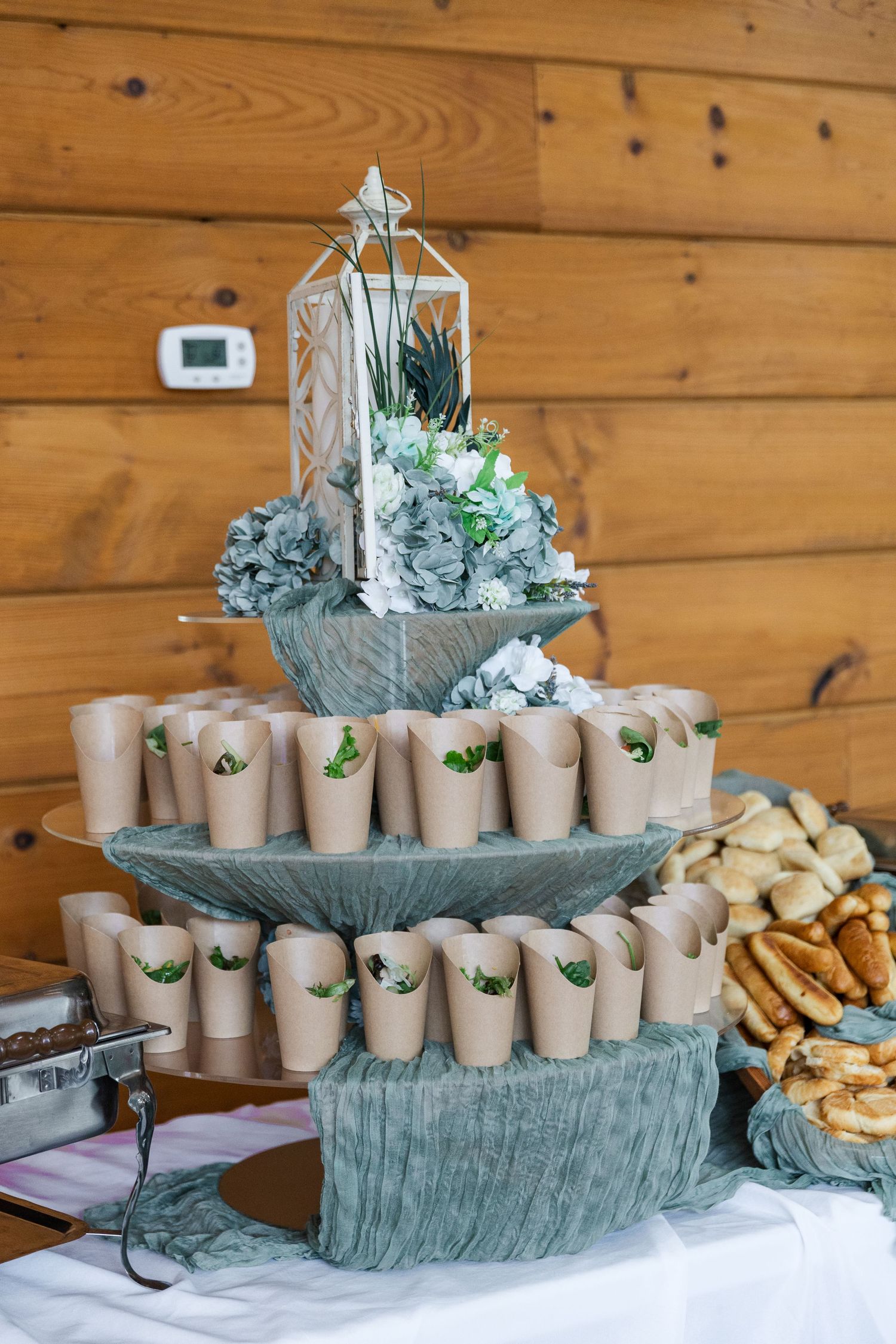 A rustic tiered serving display with paper cups and decorative lanterns arranged on wooden tables with sage green linens.