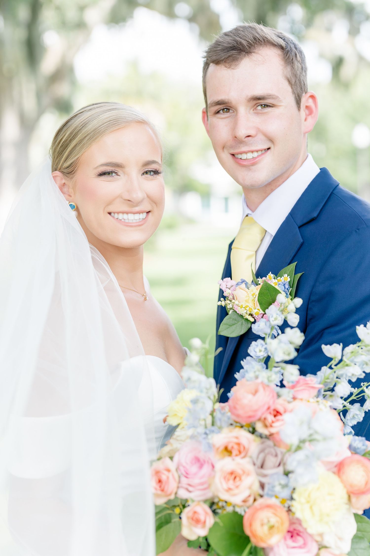 Wedding couple poses outdoors with coral and white floral bouquet against blurred garden background.