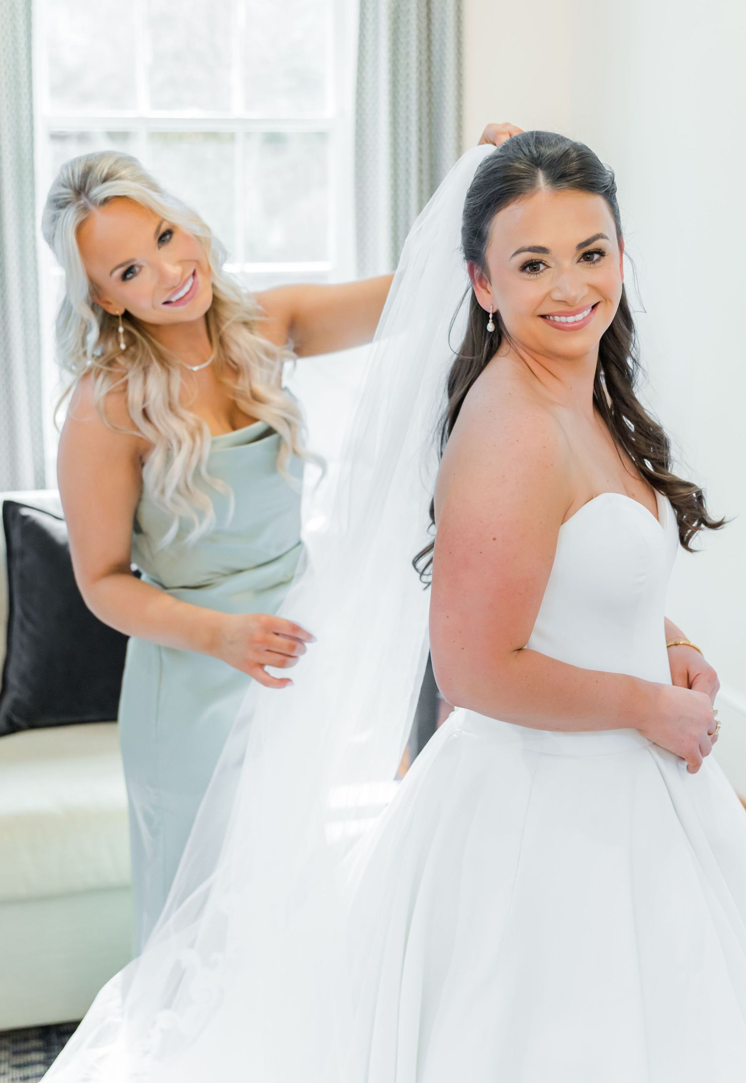Two people in formal attire embrace and smile joyfully in a bright, window-lit room.