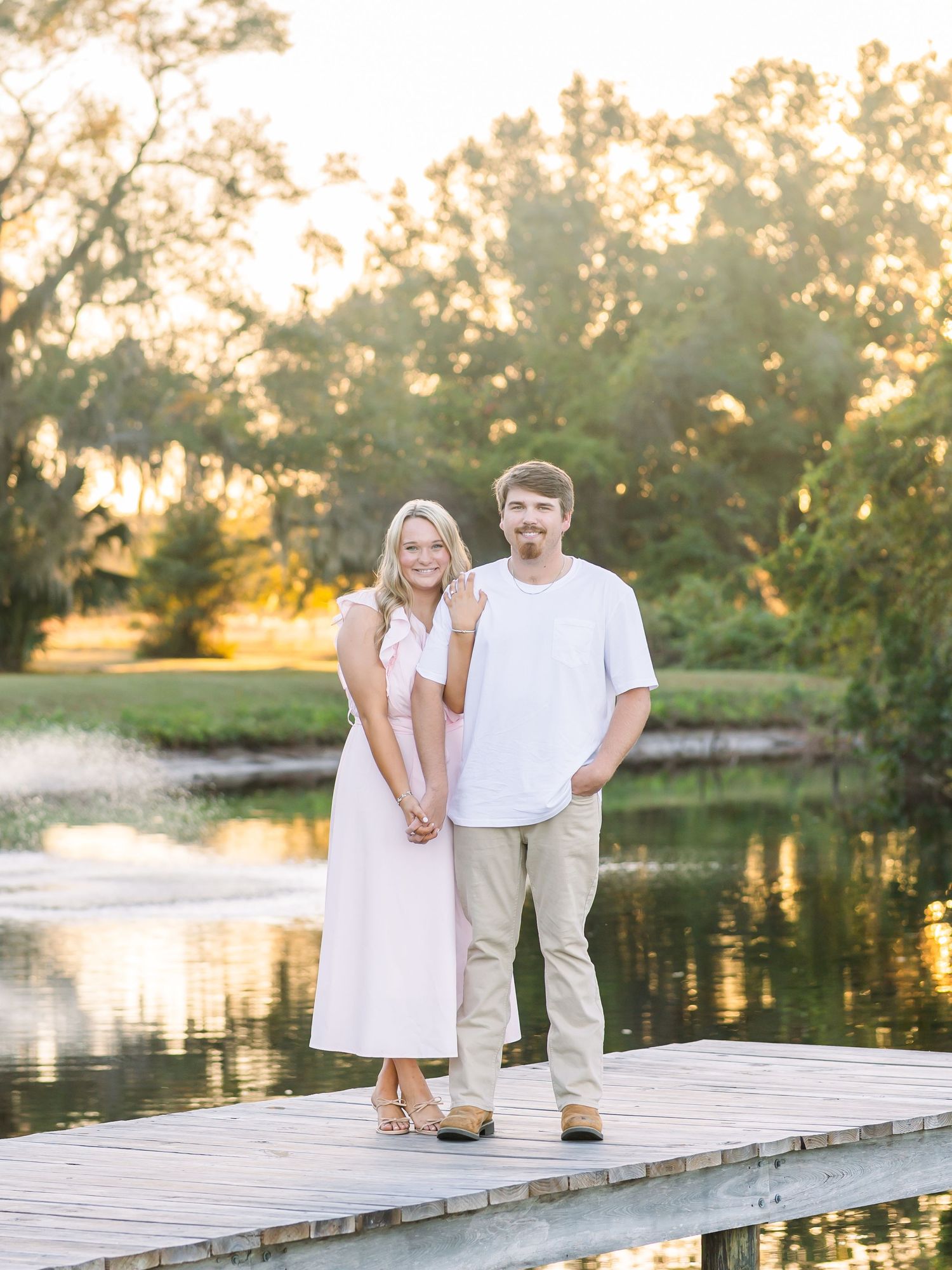 Couple in light summer attire posing together on a wooden dock at sunset by a peaceful lake.