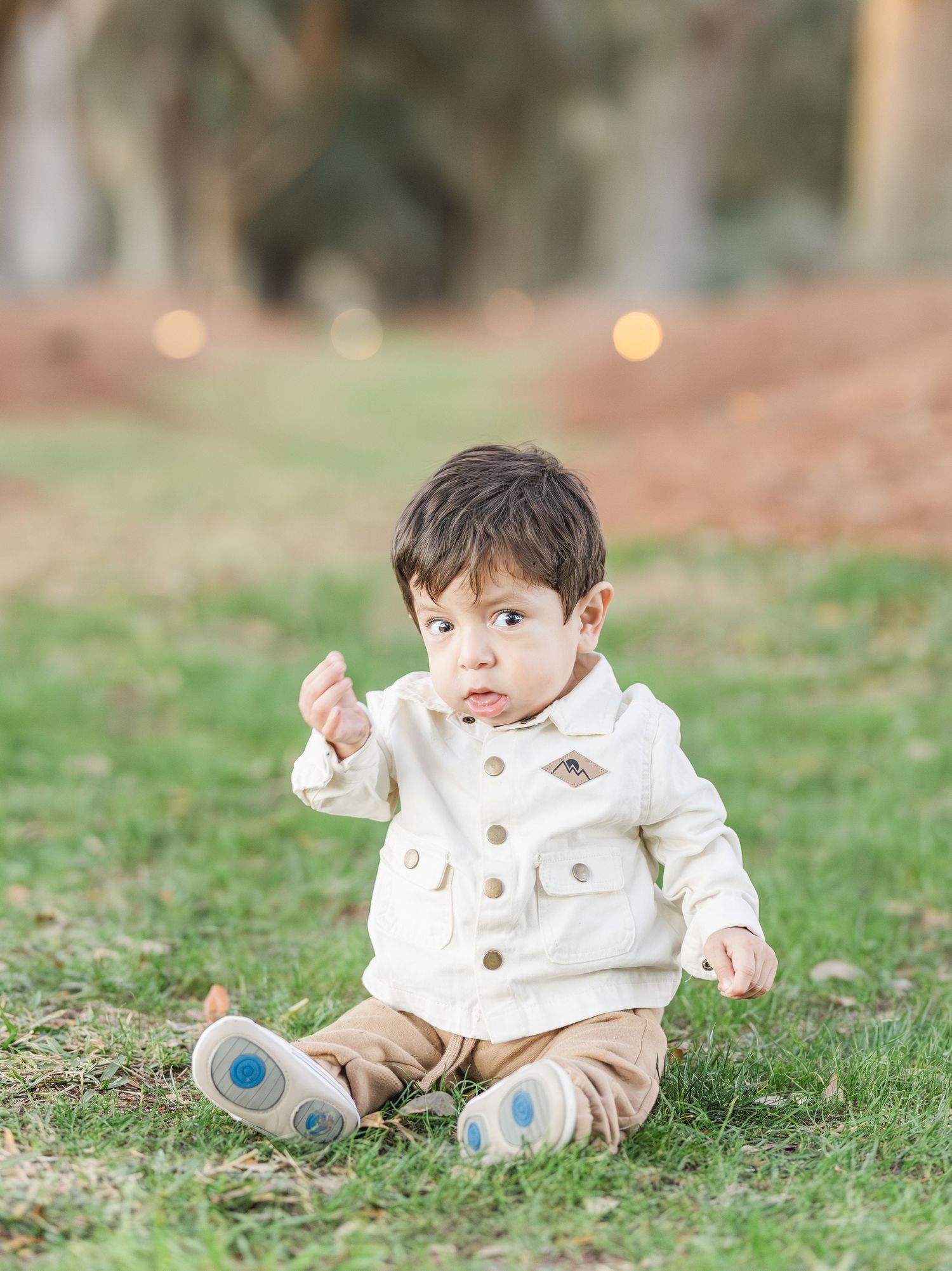 Baby in white outfit sitting on grass during outdoor photo session.