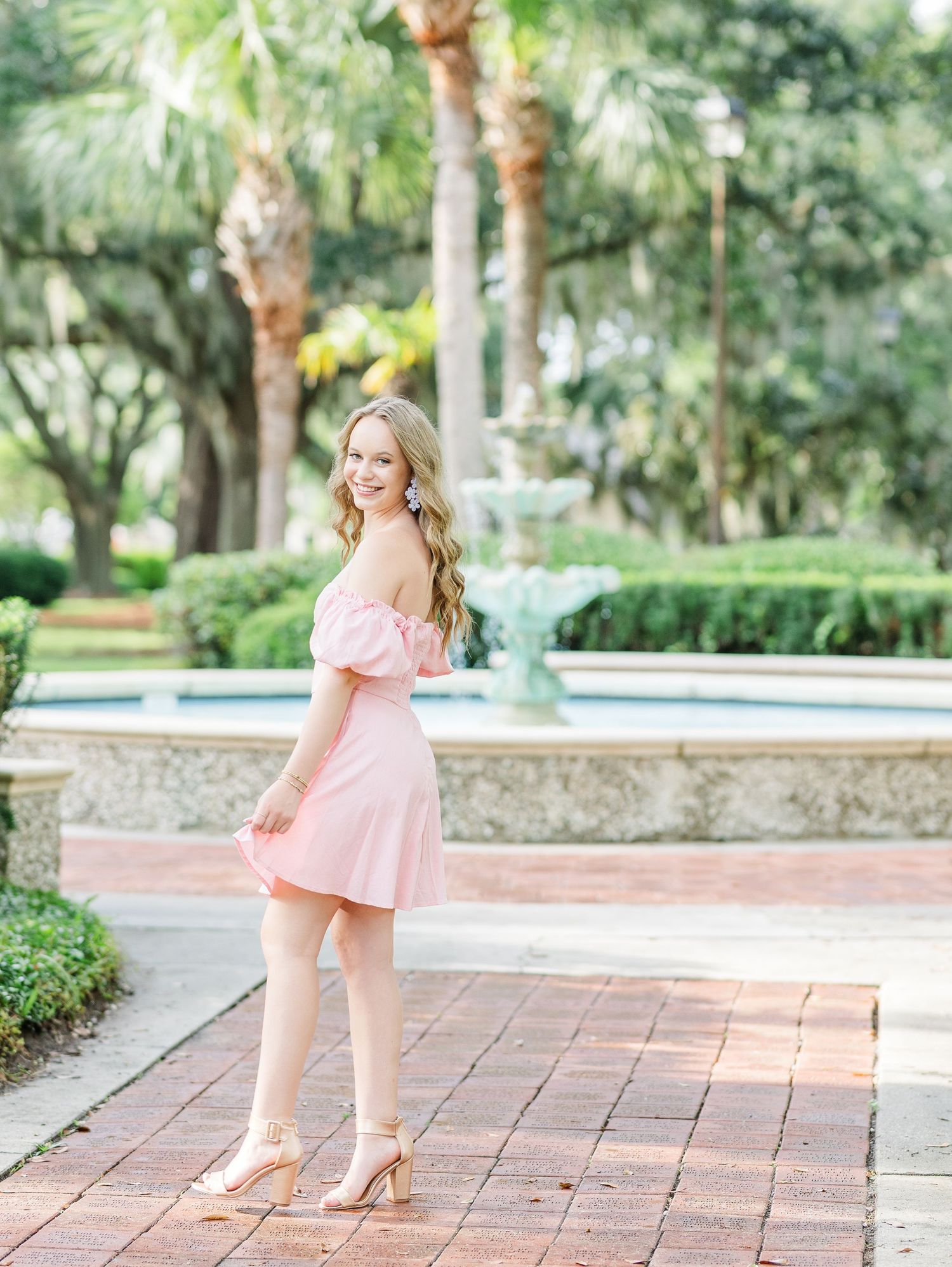 A person in a pastel pink off-shoulder dress poses on a brick pathway surrounded by tropical landscaping and palm trees.